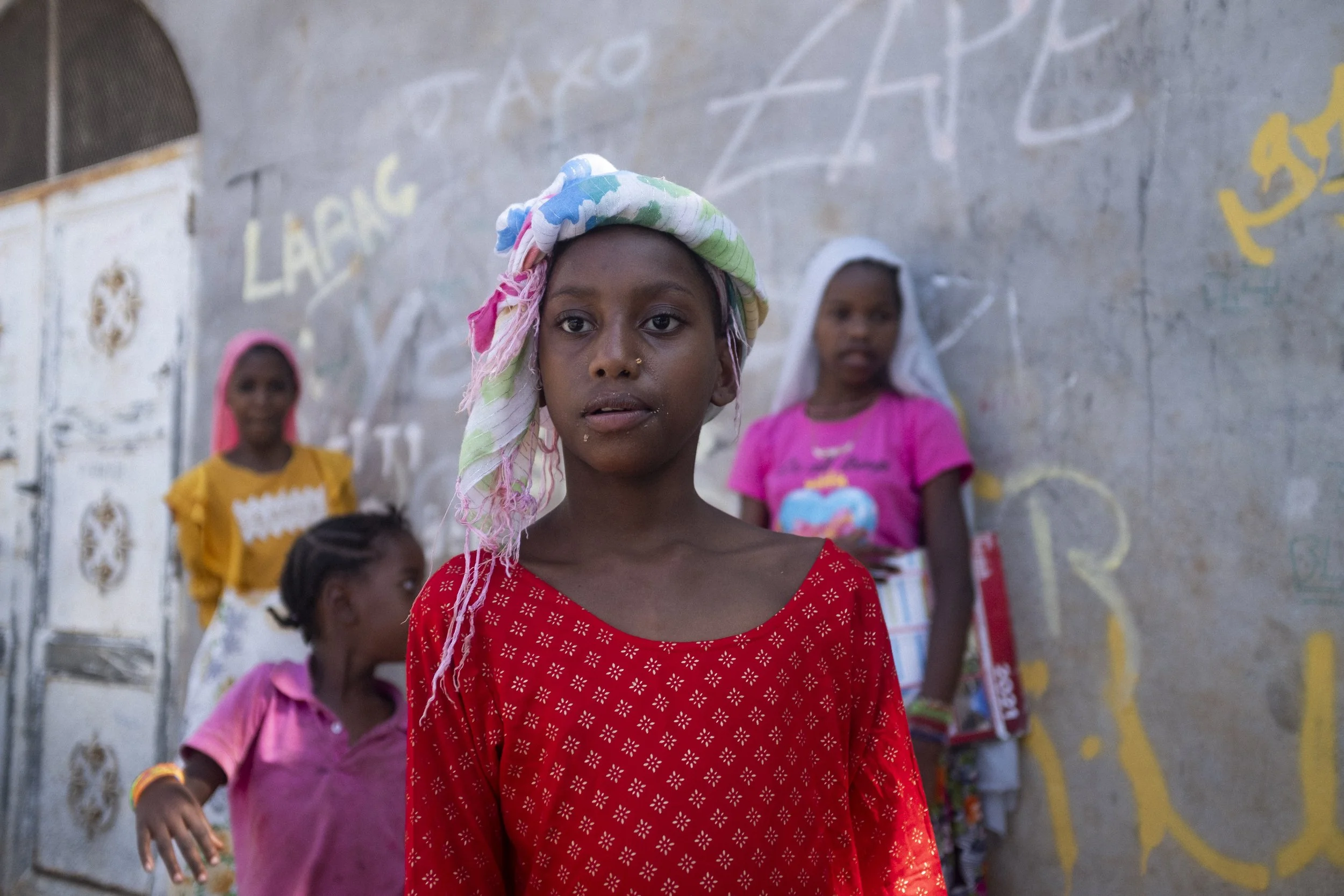 Jeune fille avec un bandeau coloré sur la tête, portant une robe rouge, devant un mur avec des dessins et des écritures, entourée d'autres filles dans un environnement extérieur.