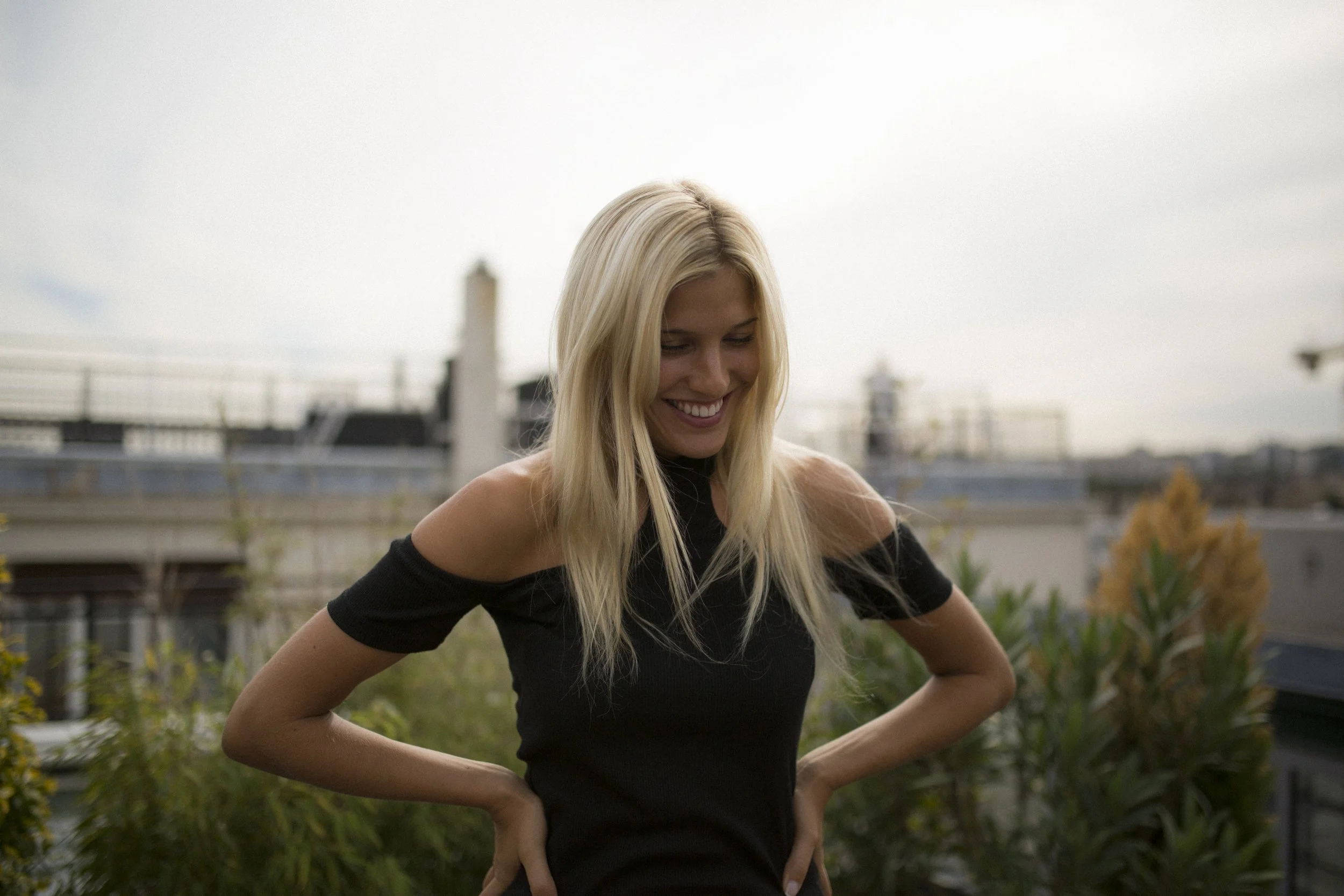 Jeune femme blonde souriante en haut noir à l'extérieur, avec des plantes et des bâtiments en arrière-plan.