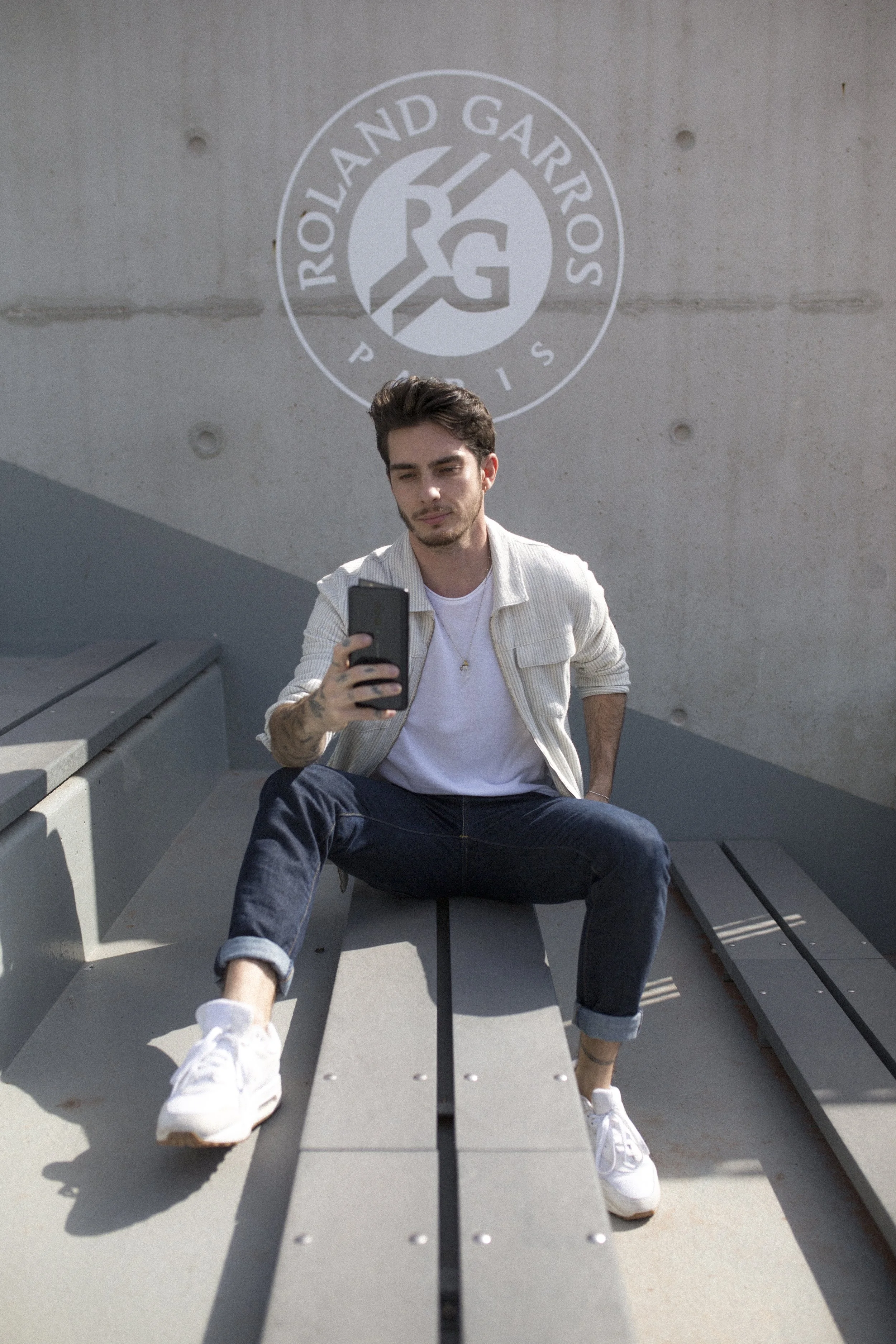 Un jeune homme assis sur un banc public, portant une veste clair, un t-shirt blanc, des jeans foncés retroussés et des baskets blanches, prenant une selfie devant un mur avec un logo du Roland-Garros.