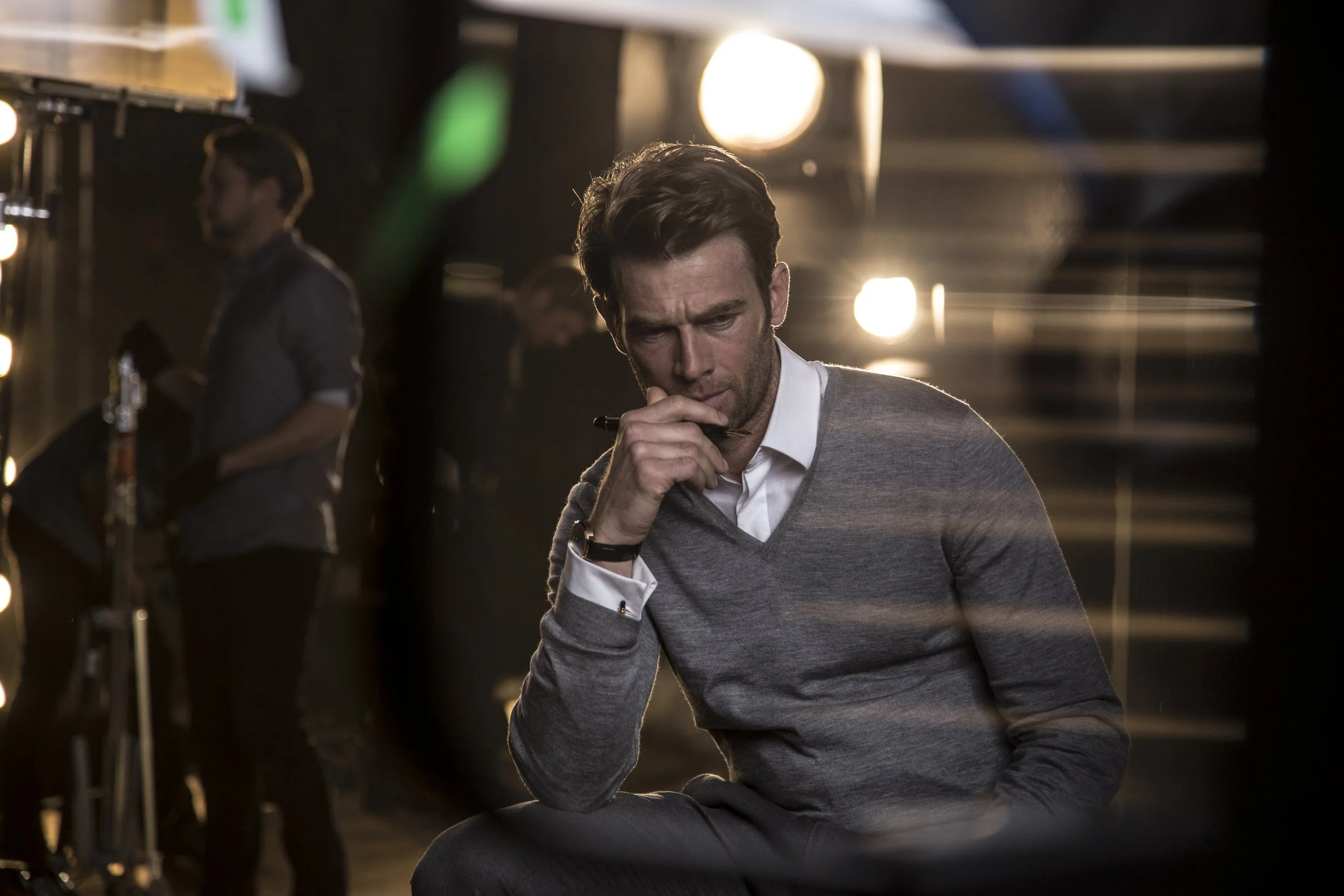 Homme en pull gris et chemise blanche assis pensif, avec une main à la bouche, dans un cadre professionnel ou créatif, avec des lumières derrière lui.