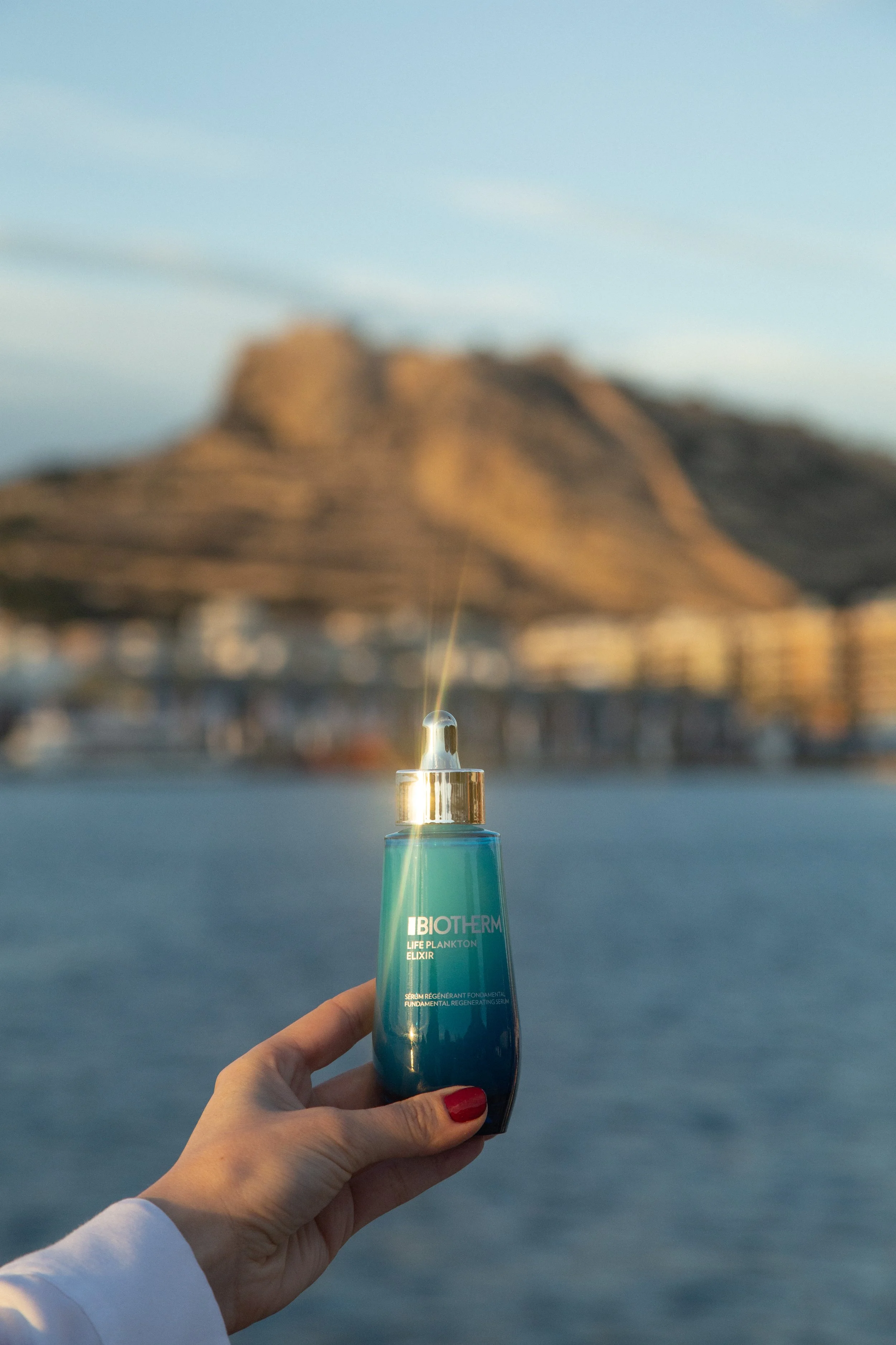Une main tenant un flacon de produit de soin Biotherm, avec une montagne et le ciel en arrière-plan.