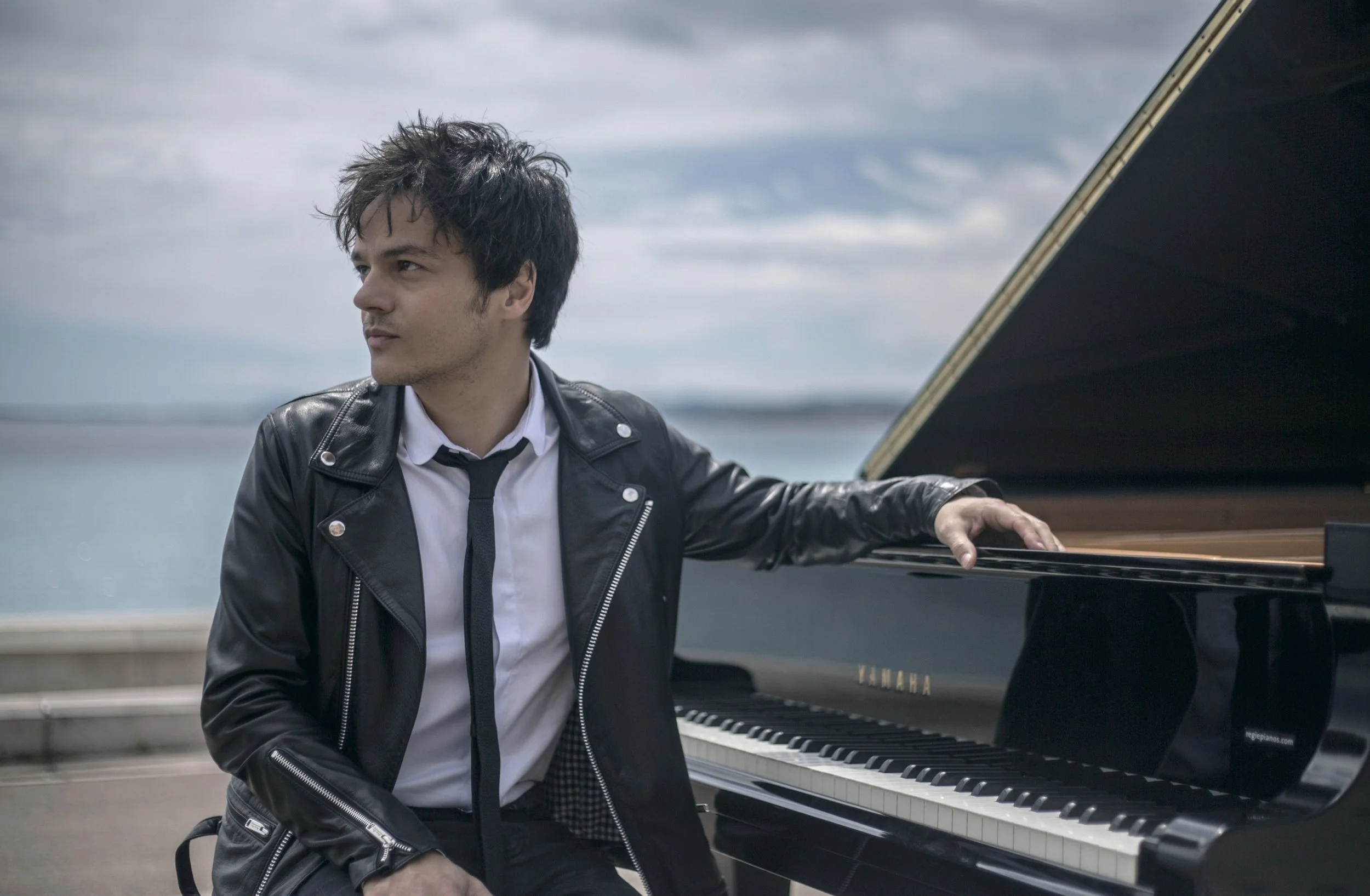 Un homme avec des cheveux noirs et ébouriffés, portant une veste en cuir noire, une chemise blanche et une cravate noire, est assis devant un piano noir Yamaha, près d'une mer ou d'un lac, avec un ciel nuageux en arrière-plan.