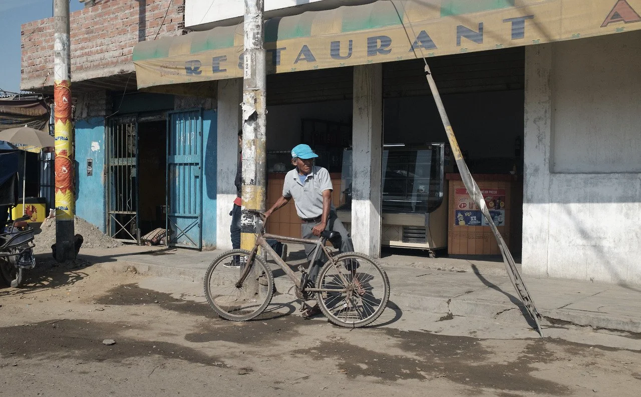 Un homme avec un chapeau bleu se tient à côté d'un vélo rouillé sur une chaussée poussiéreuse devant une devanture de restaurant en construction ou en rénovation, avec un panneau frontal de restaurant partiellement visible.