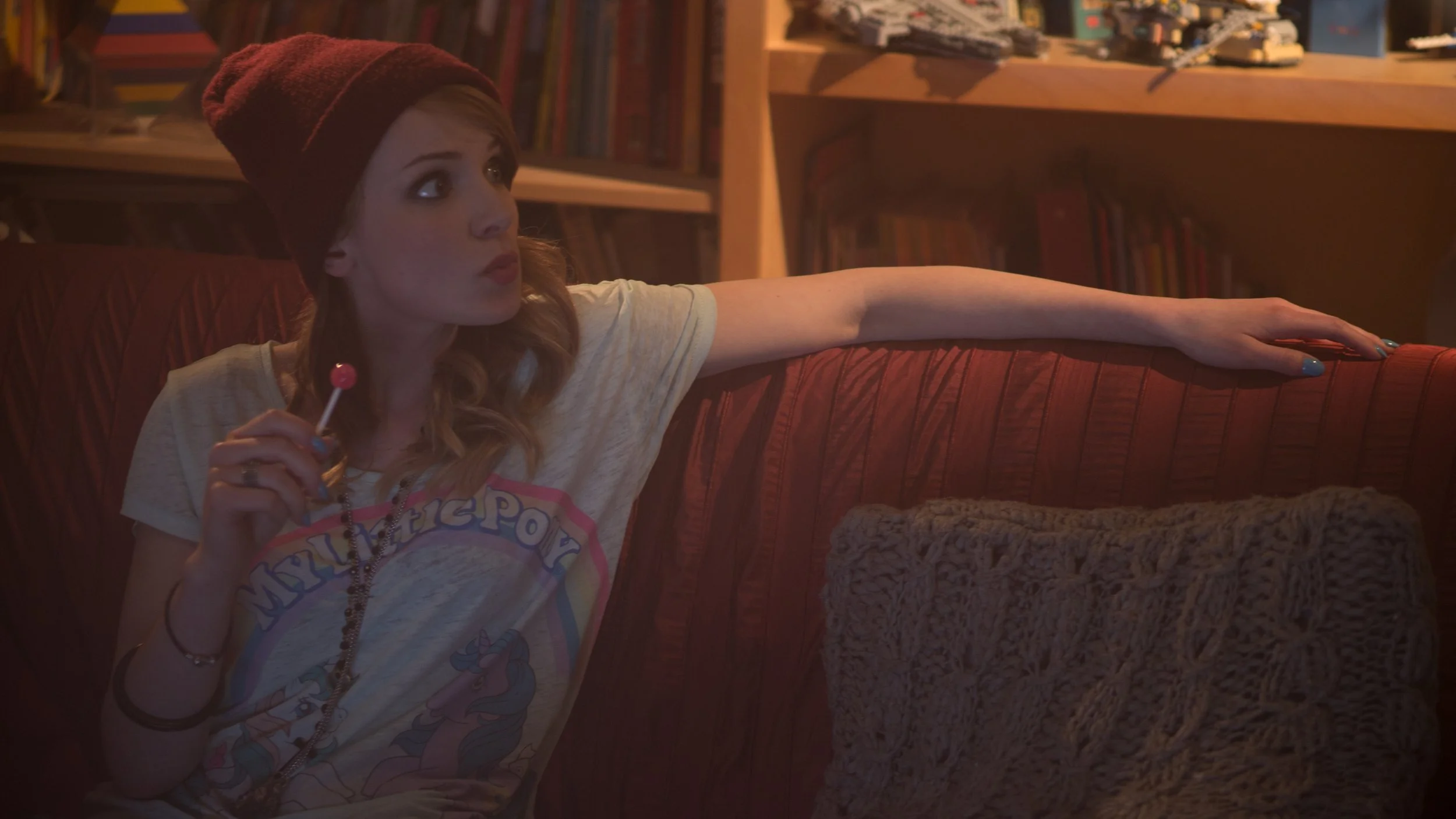 Une jeune femme assise sur un canapé rouge, portant un t-shirt avec le logo My Little Pony, une casquette rouge, en tenant une sucette. La pièce est éclairée de manière chaleureuse, avec une bibliothèque en arrière-plan.