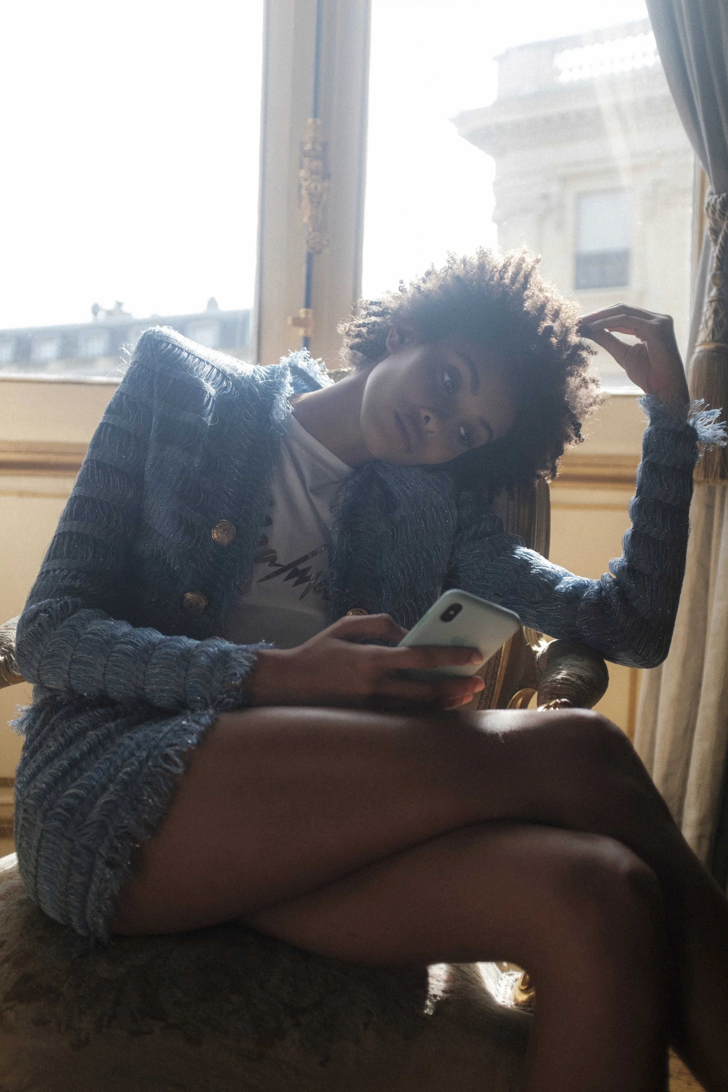 Jeune femme assise sur un fauteuil en cuir, tenant un téléphone portable, dans une pièce avec une grande fenêtre laissant entrer la lumière naturelle.