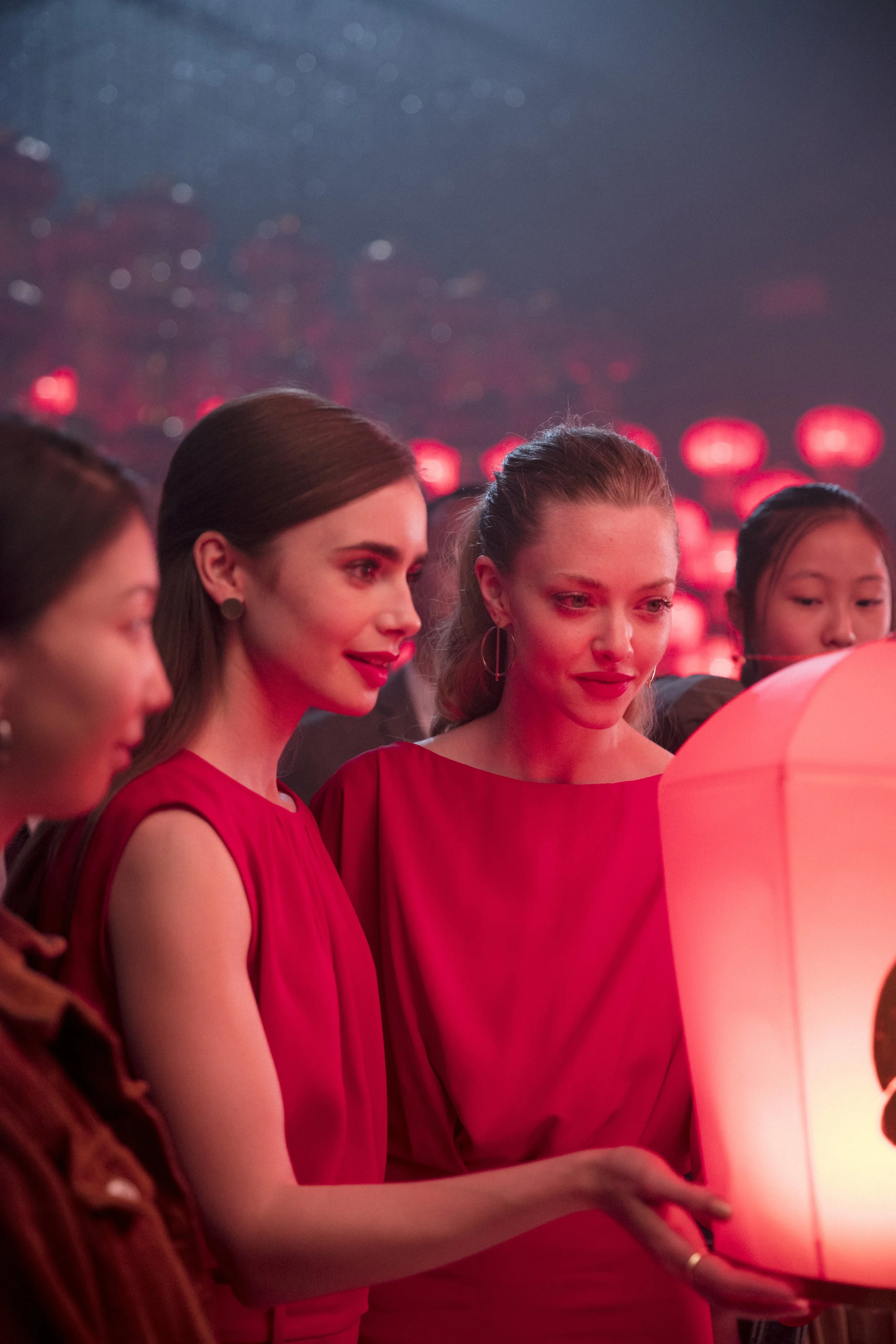 Groupe de femmes vêtues de rouge regardant une lanterne en papier lors d'une célébration nocturne avec des lumières rouges en arrière-plan.