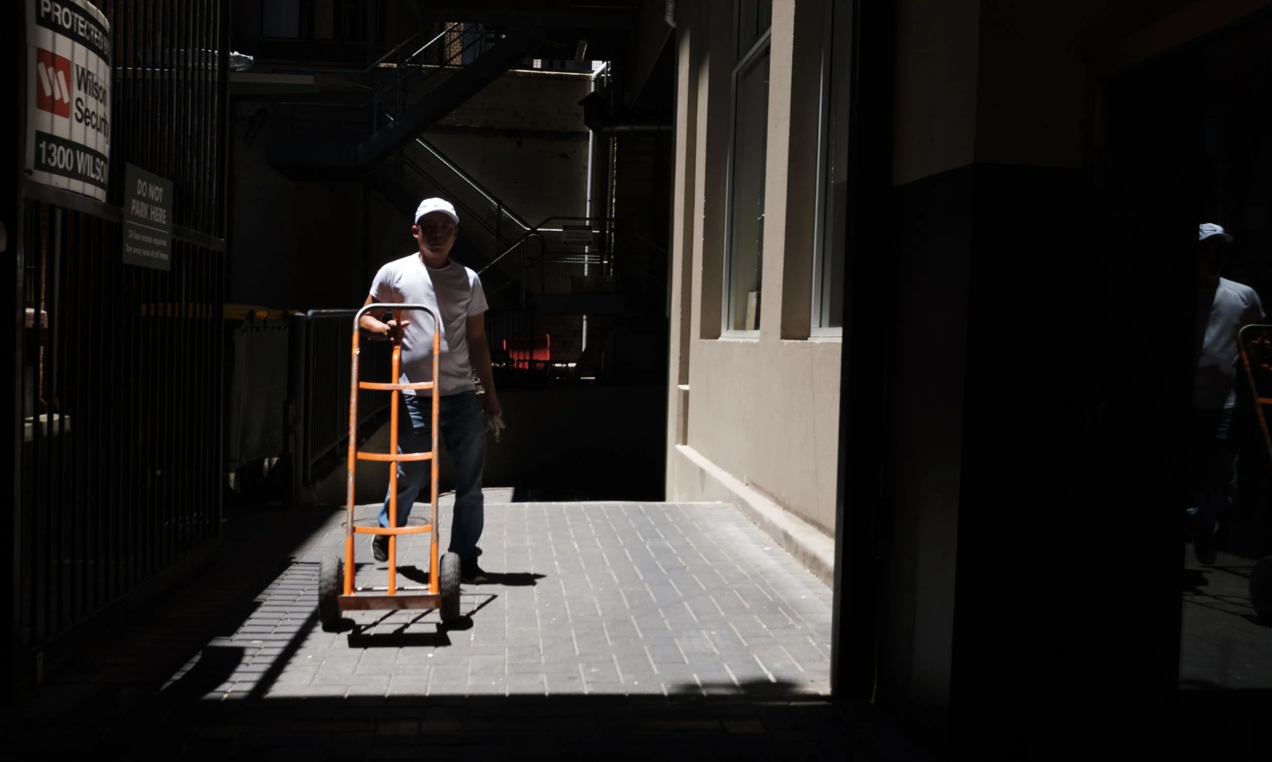 Un homme portant une casquette blanche pousse un chariot orange dans une zone sombre, en dehors d'un bâtiment avec entrée ensoleillée
