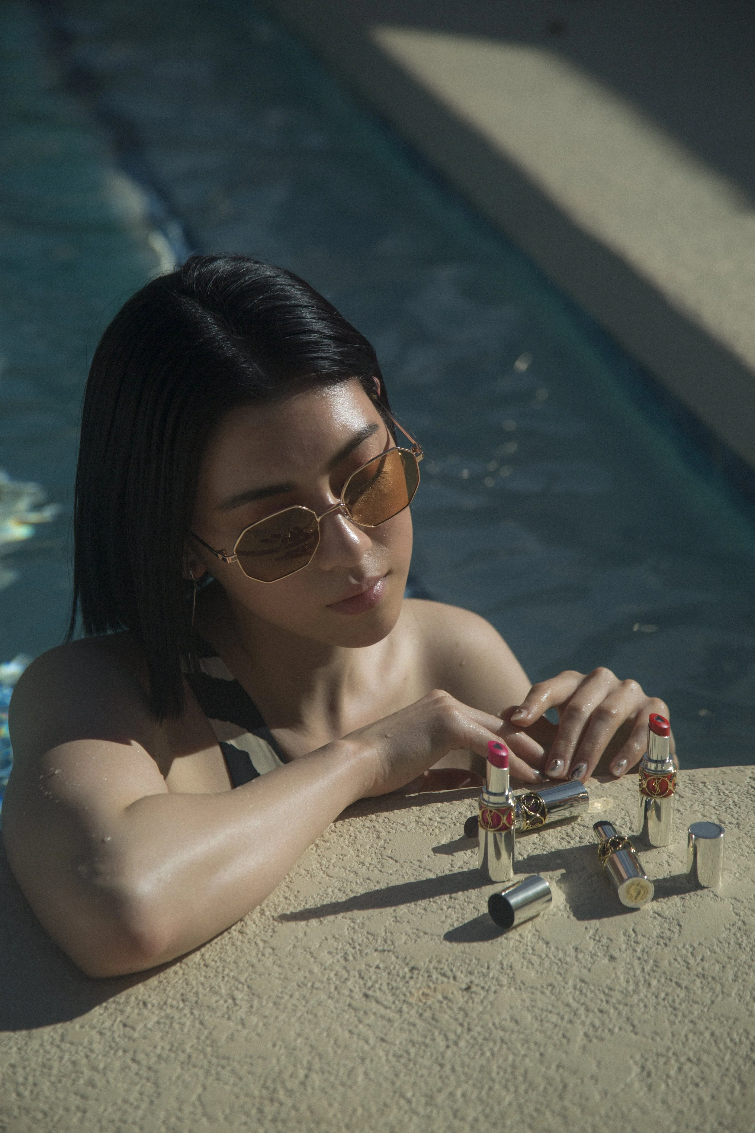Une femme nageant dans une piscine en portant des lunettes de soleil, en train d'examiner ou de mettre du rouge à lèvres sur le rebord de la piscine.