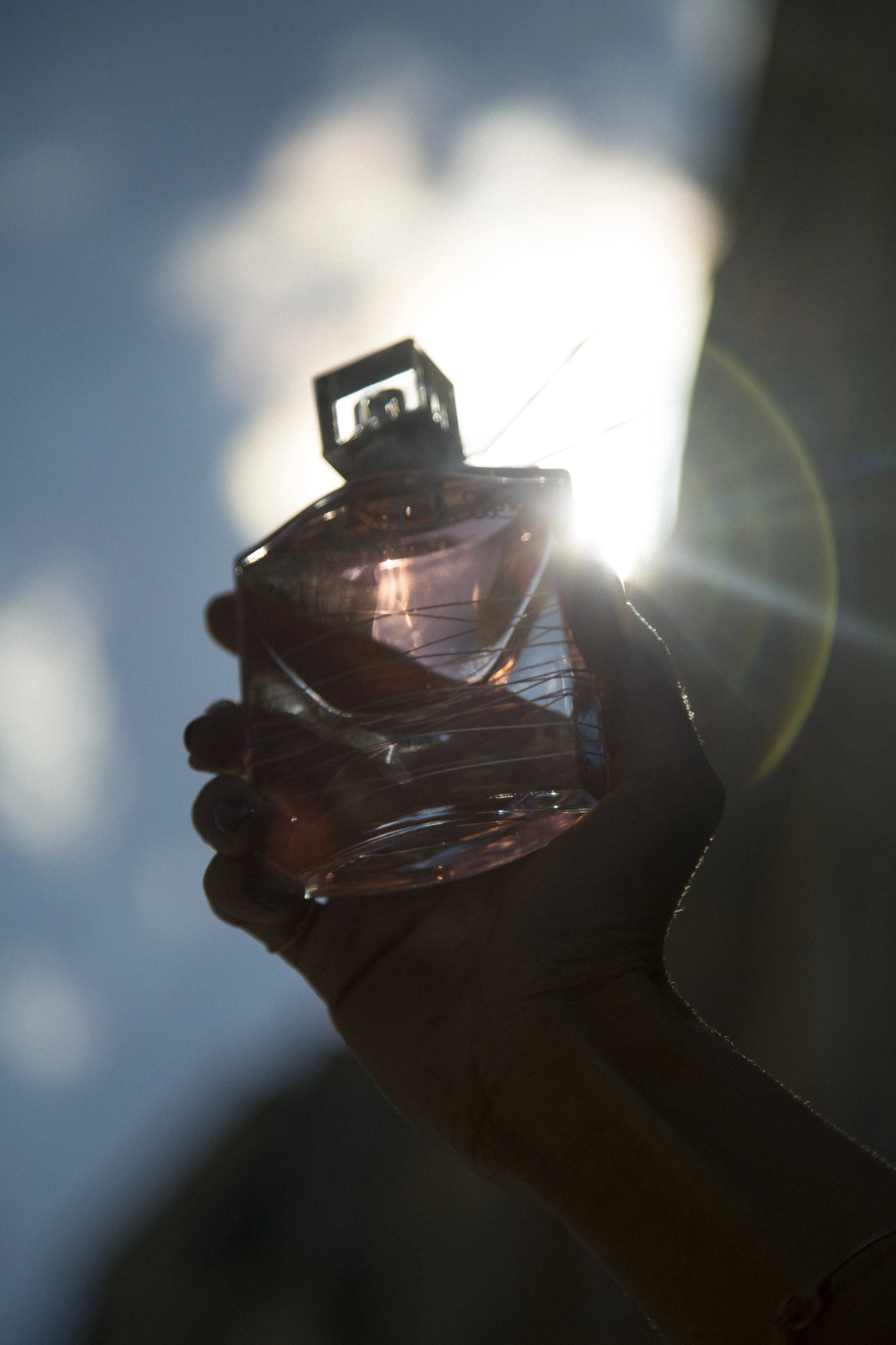 Une main tenant un flacon de parfum contre le soleil, créant un reflet de lumière et un halo lumineux.
