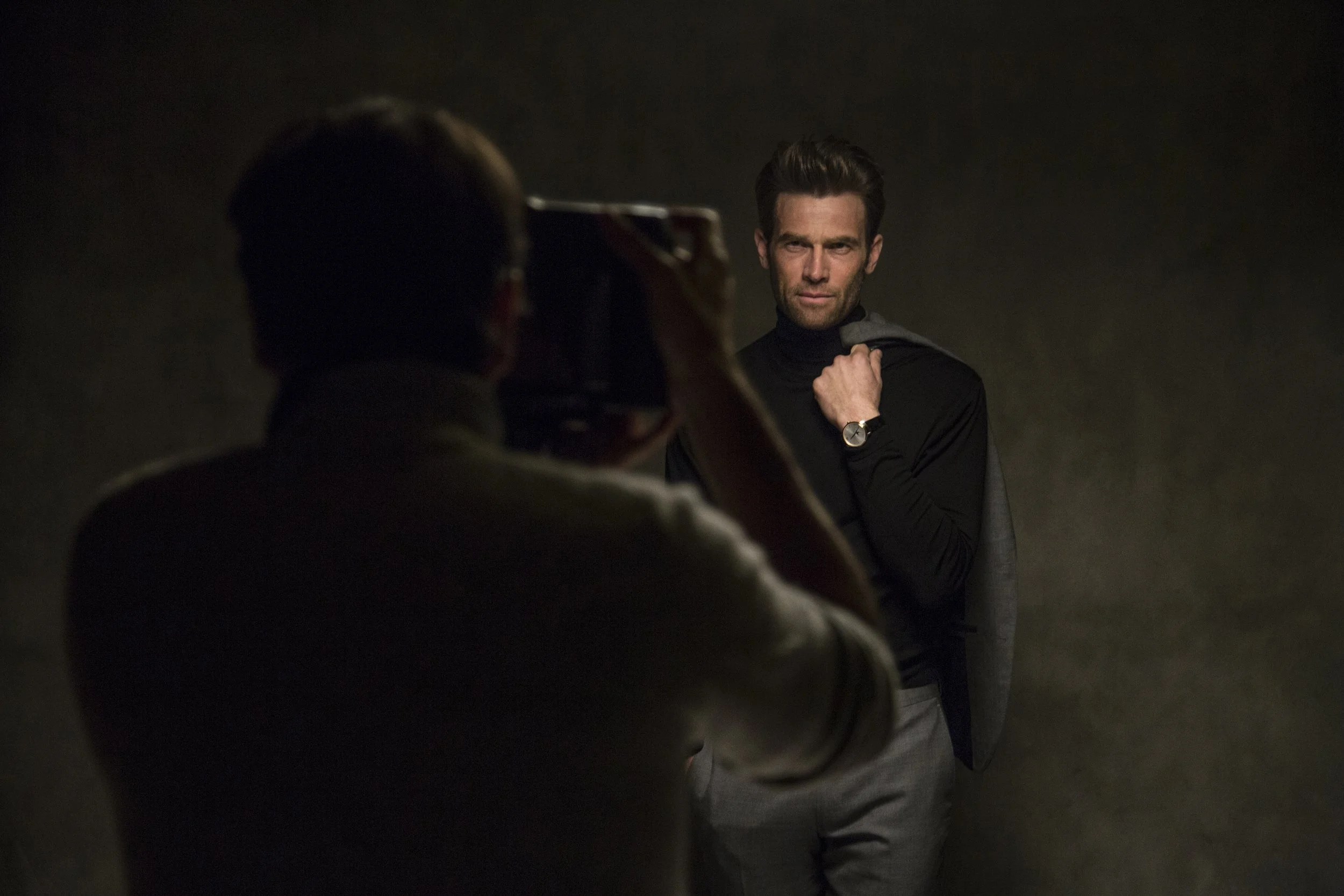 Un homme posant pour une séance photo, assis dans une pièce sombre, portant un pull noir et une montre au poignet gauche, regardant sérieusement l'appareil photo.