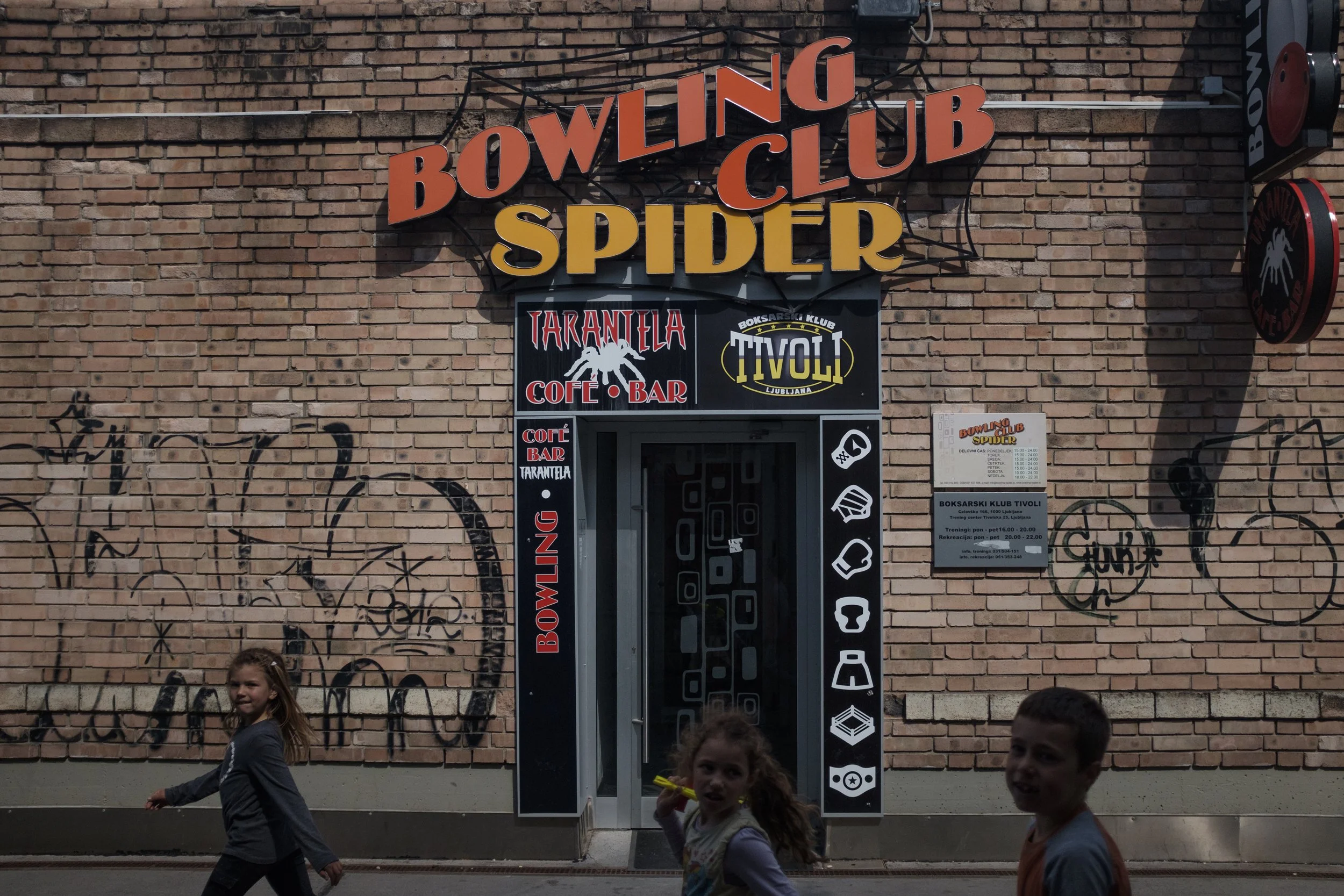 Entrée du club de bowling 'Spider' avec un mur en briques derrière, des graffitis, des affiches et des enfants qui passent devant.