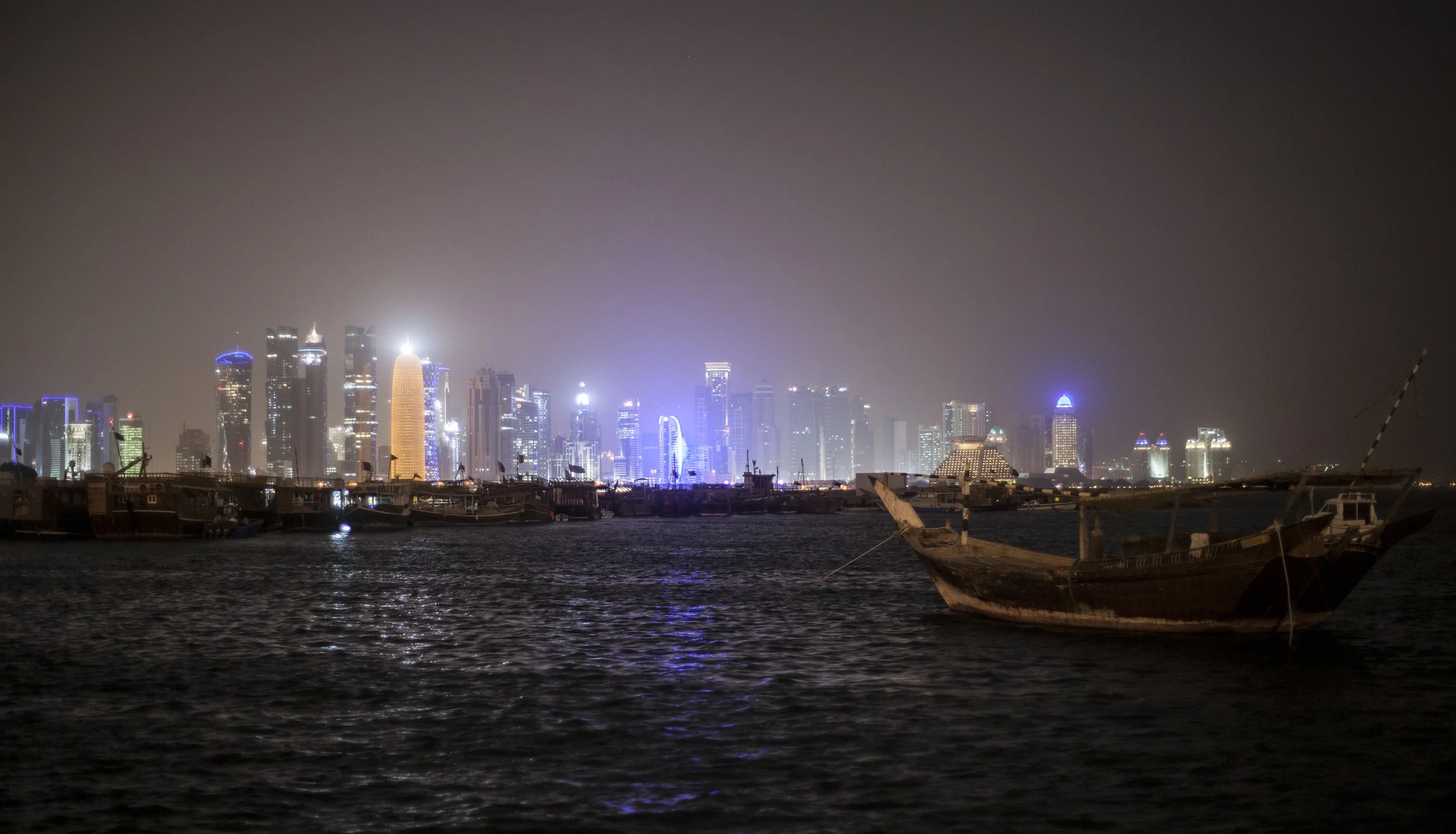 Une vue de la ligne d'horizon de la ville de Doha illuminée la nuit, avec des bâtiments modernes et des gratte-ciel, vue depuis la mer avec un vieux bateau en bois en premier plan.