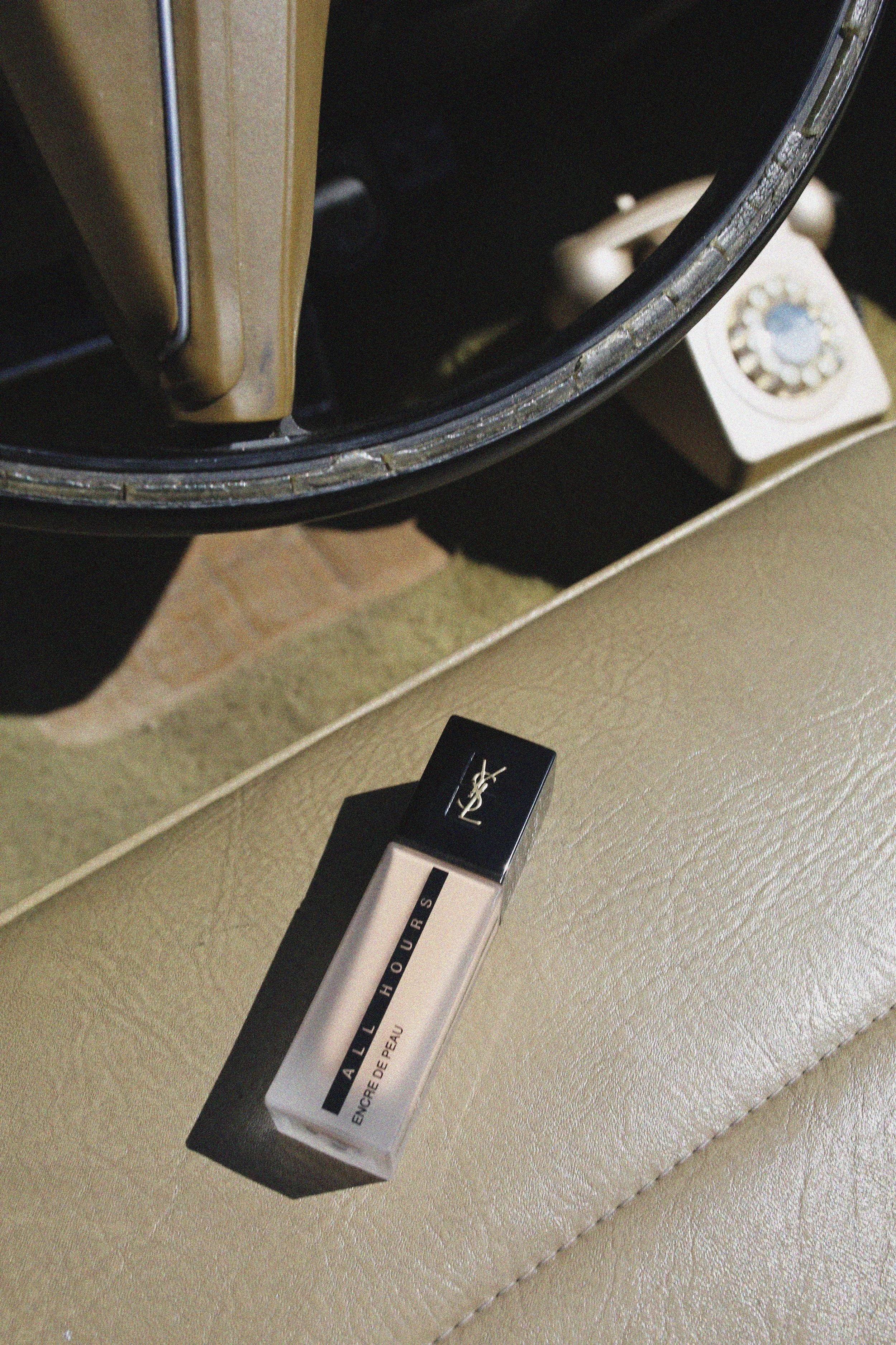 Un tube de rouge à lèvres Yves Saint Laurent All Hours repose sur une surface en cuir beige, avec un tableau de bord en arrière-plan.