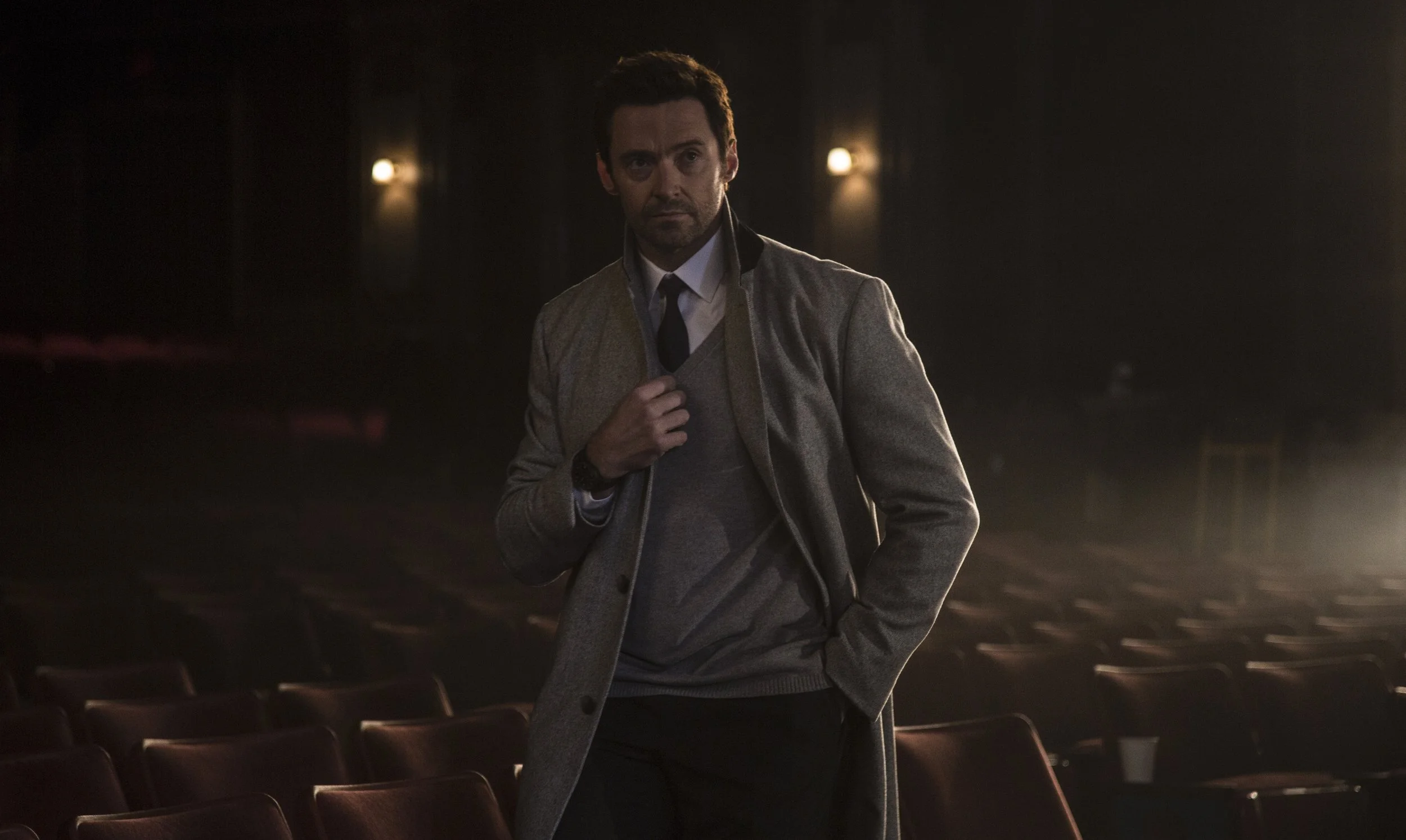 Un homme en manteau gris, chemise blanche et cravate dans une salle de théâtre sombre, avec des sièges en avant-plan.