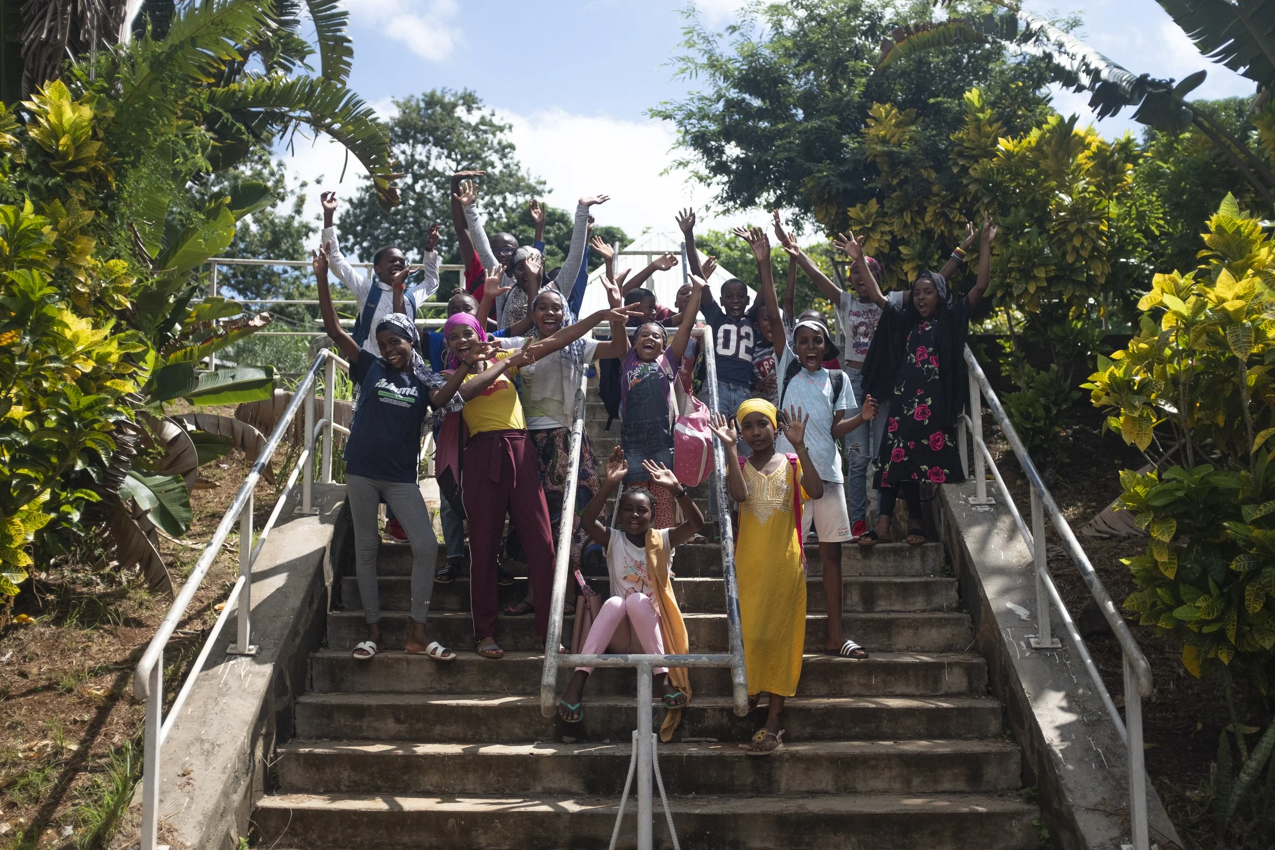 Groupe d'enfants souriants et joyeux montant les escaliers en extérieur, entourés de verdure et sous un ciel ensoleillé.