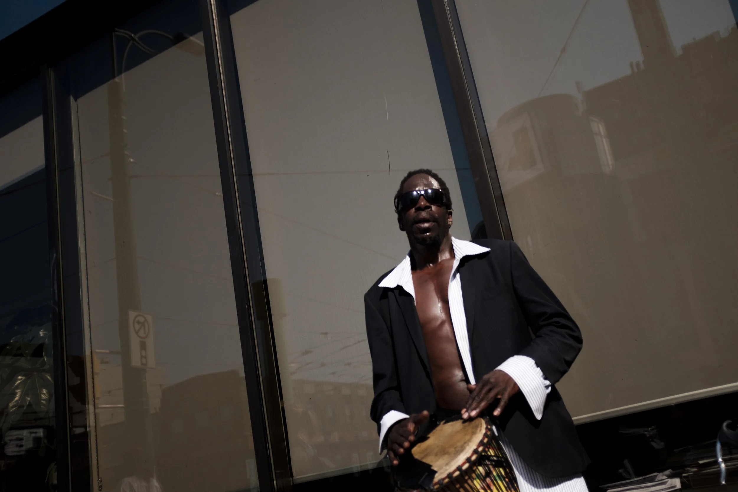 Un homme noir portant des lunettes de soleil, une chemise ouverte et un costume noir, joue d'un instrument de percussion devant une façade en verre reflétant le ciel et des bâtiments.