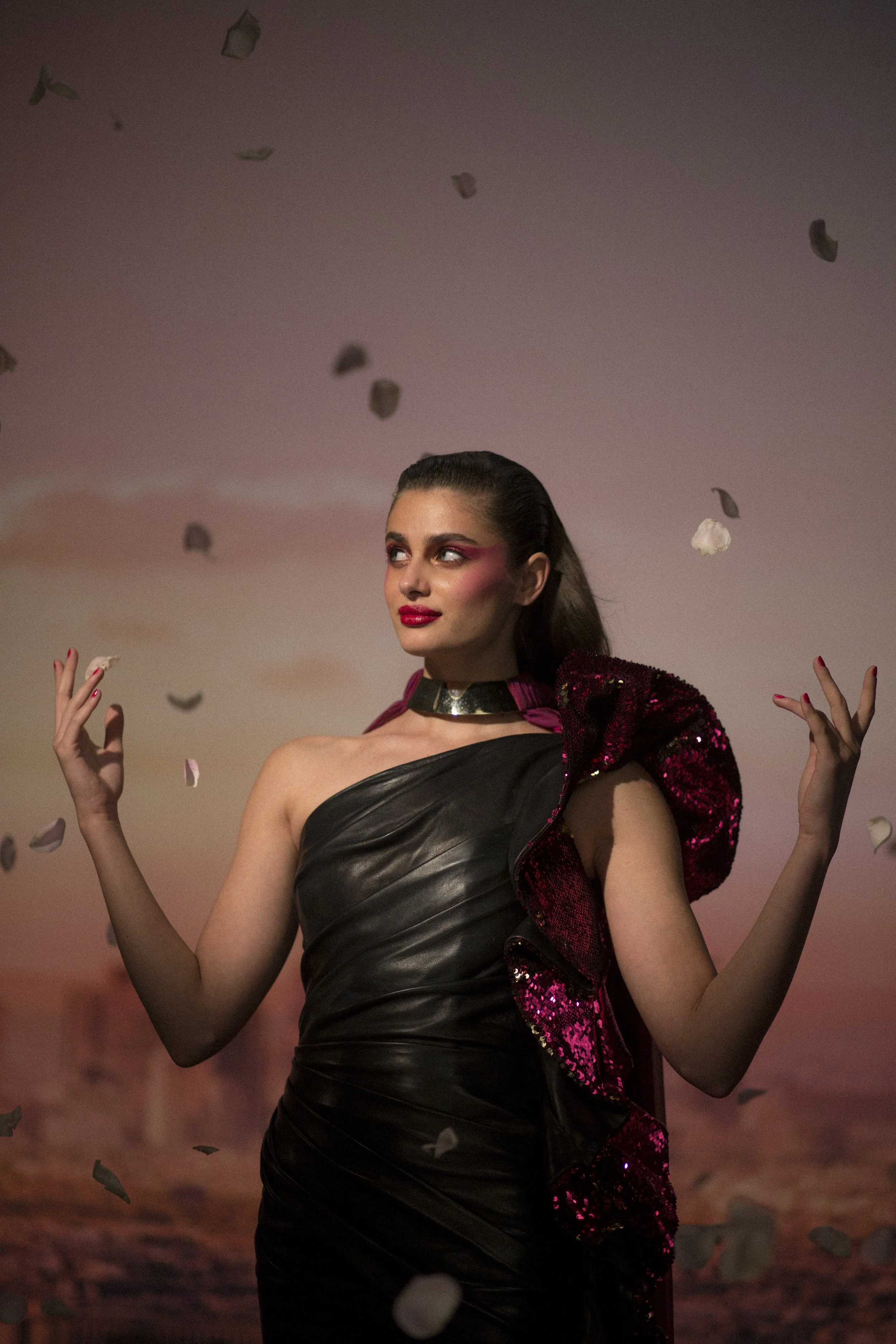 Une femme élégante portant une robe noire en cuir avec un accessoire en sequins rose foncé, se tenant sous un ciel rose vaporeux avec des pétales qui tombent.