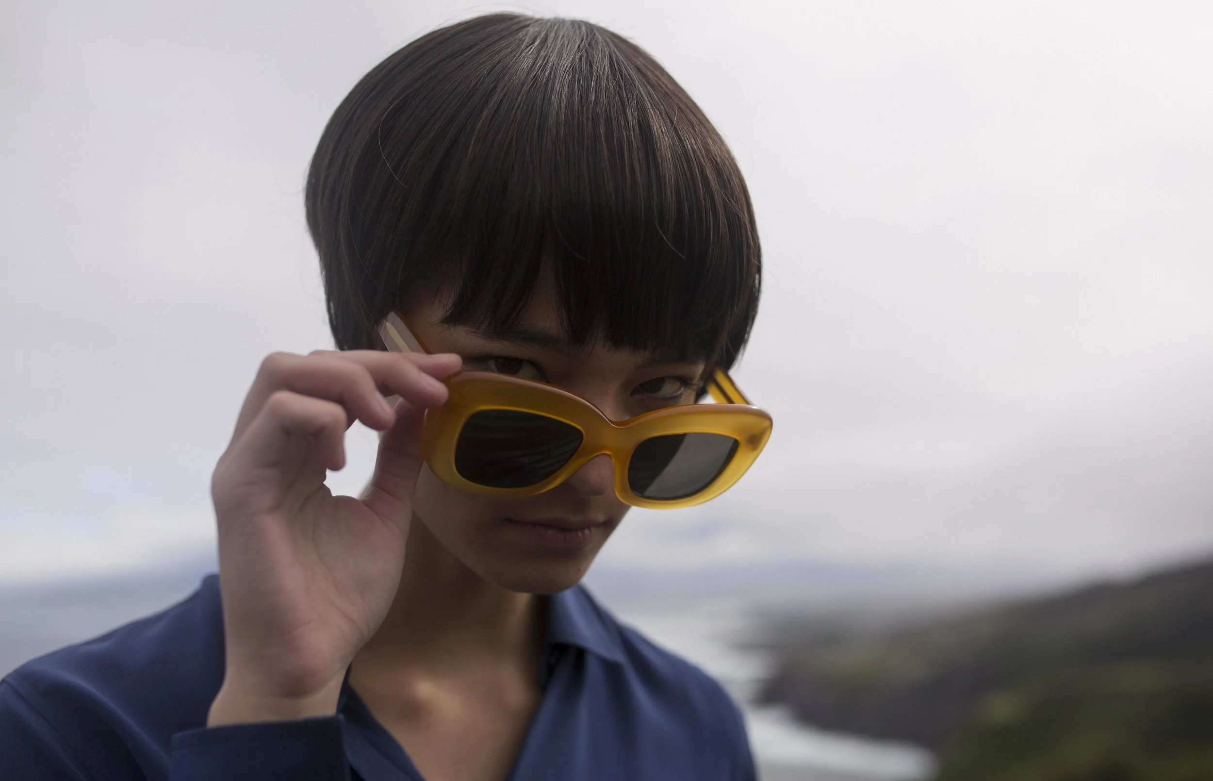 Une personne portant des lunettes de soleil jaunes regarde par-dessus ses lunettes dans un décor extérieur avec une mer ou un océan et une côte à l'arrière-plan, sous un ciel nuageux.