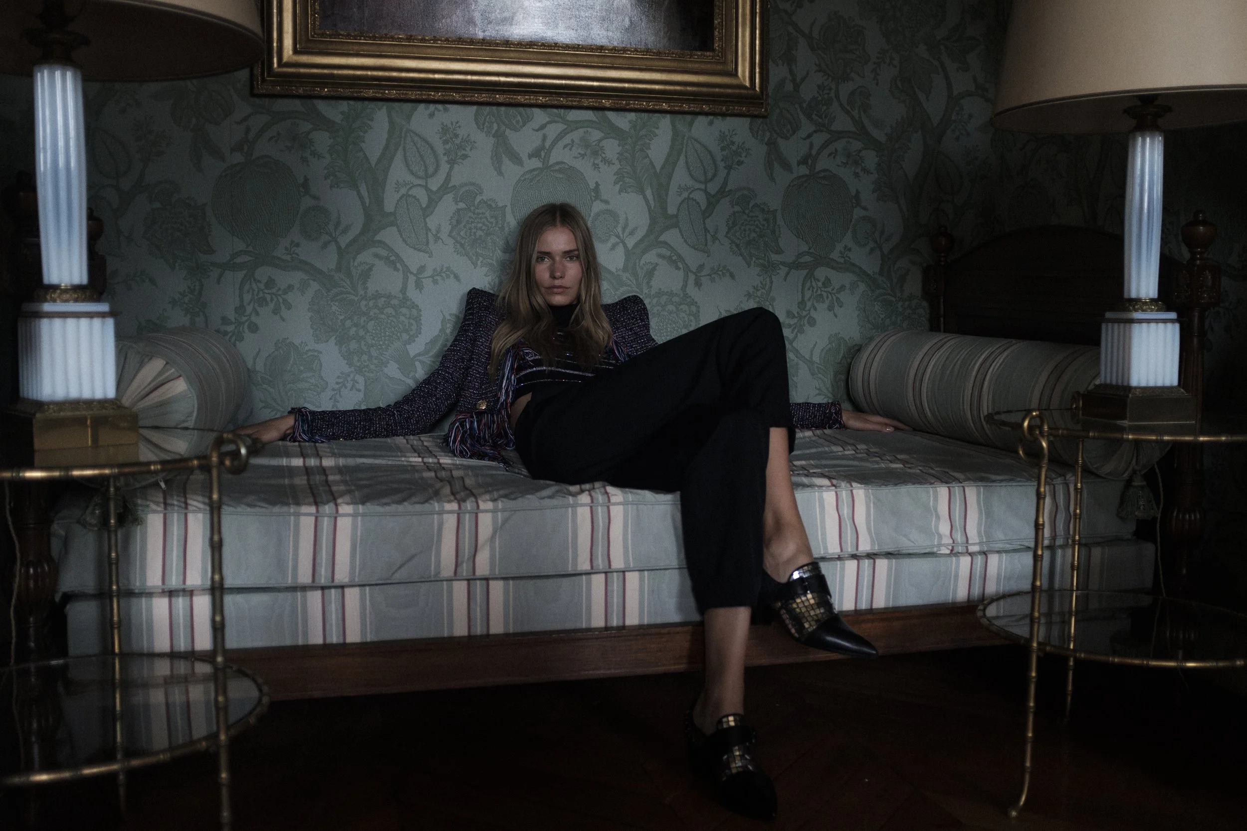 Une jeune femme assise sur un lit dans une chambre décorée de papier peint floral, portant une tenue noire et un blazer à textures, avec des chaussures à talons avec des détails en or.