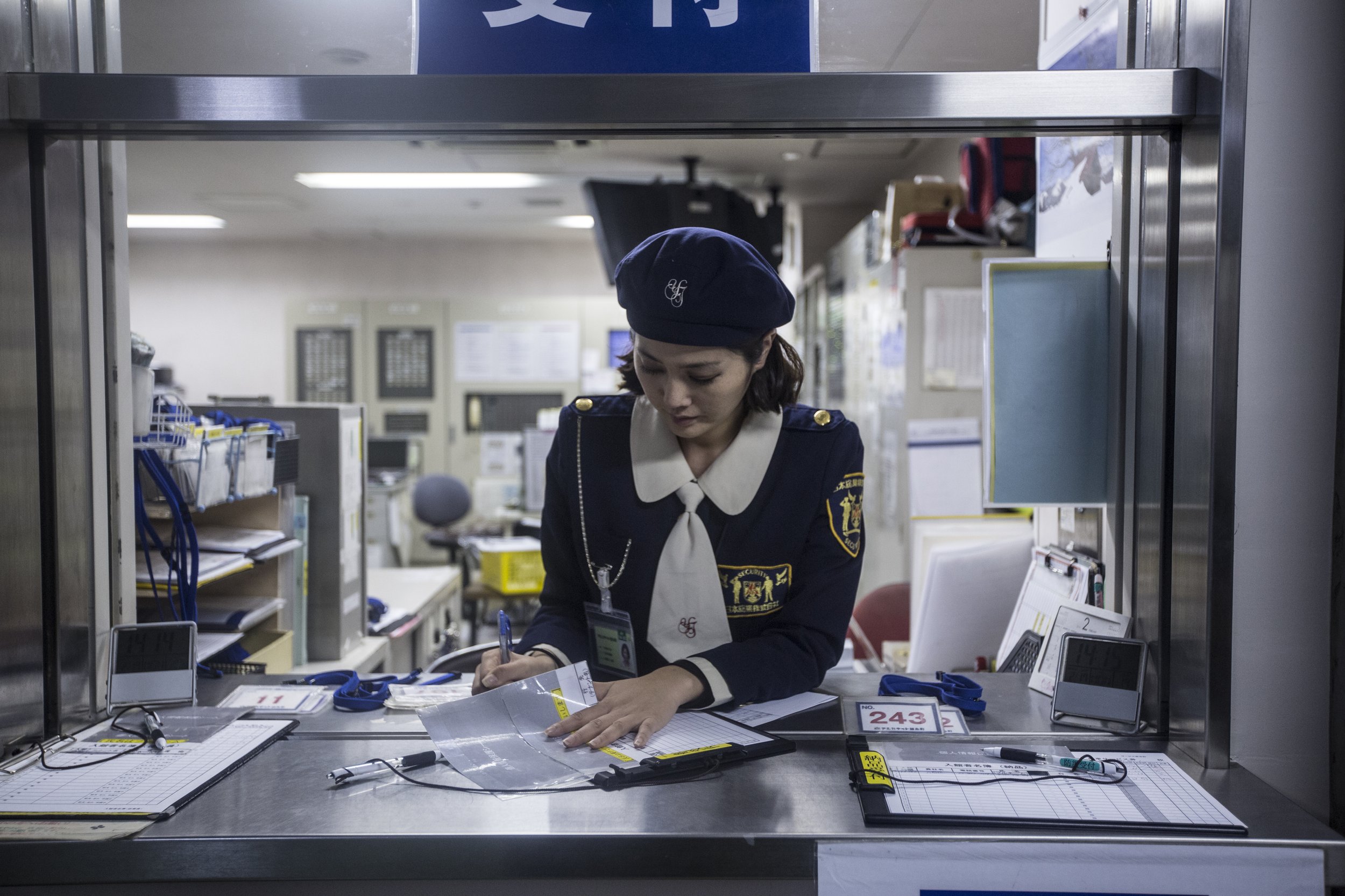 Une femme en uniforme de bureau de police ou de sécurité à un poste de contrôle, vérifiant des documents avec un stylo. L'environnement est un bureau ou centre de services avec des papiers, un ordinateur, des horodateurs, et du matériel de bureau.