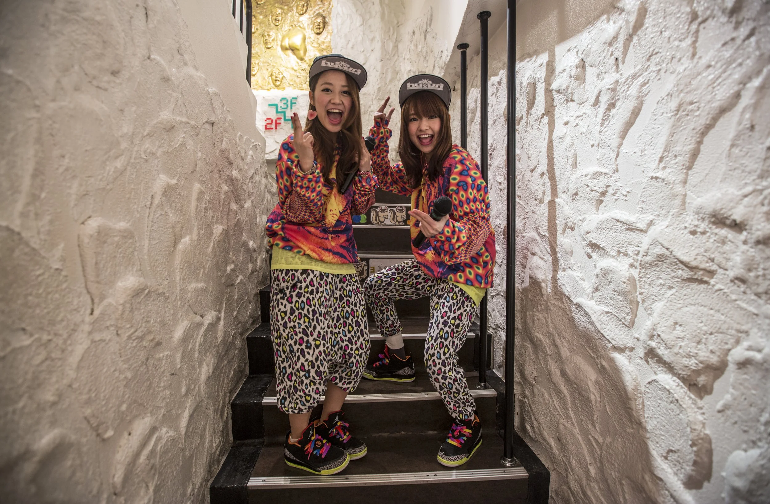 Deux jeunes femmes souriantes posant sur un escalier en intérieur, avec murs blancs en pierre, portant des vêtements colorés à motifs léopard et polka dots, toutes deux portant des casquettes et des chaussures de sport colorées.