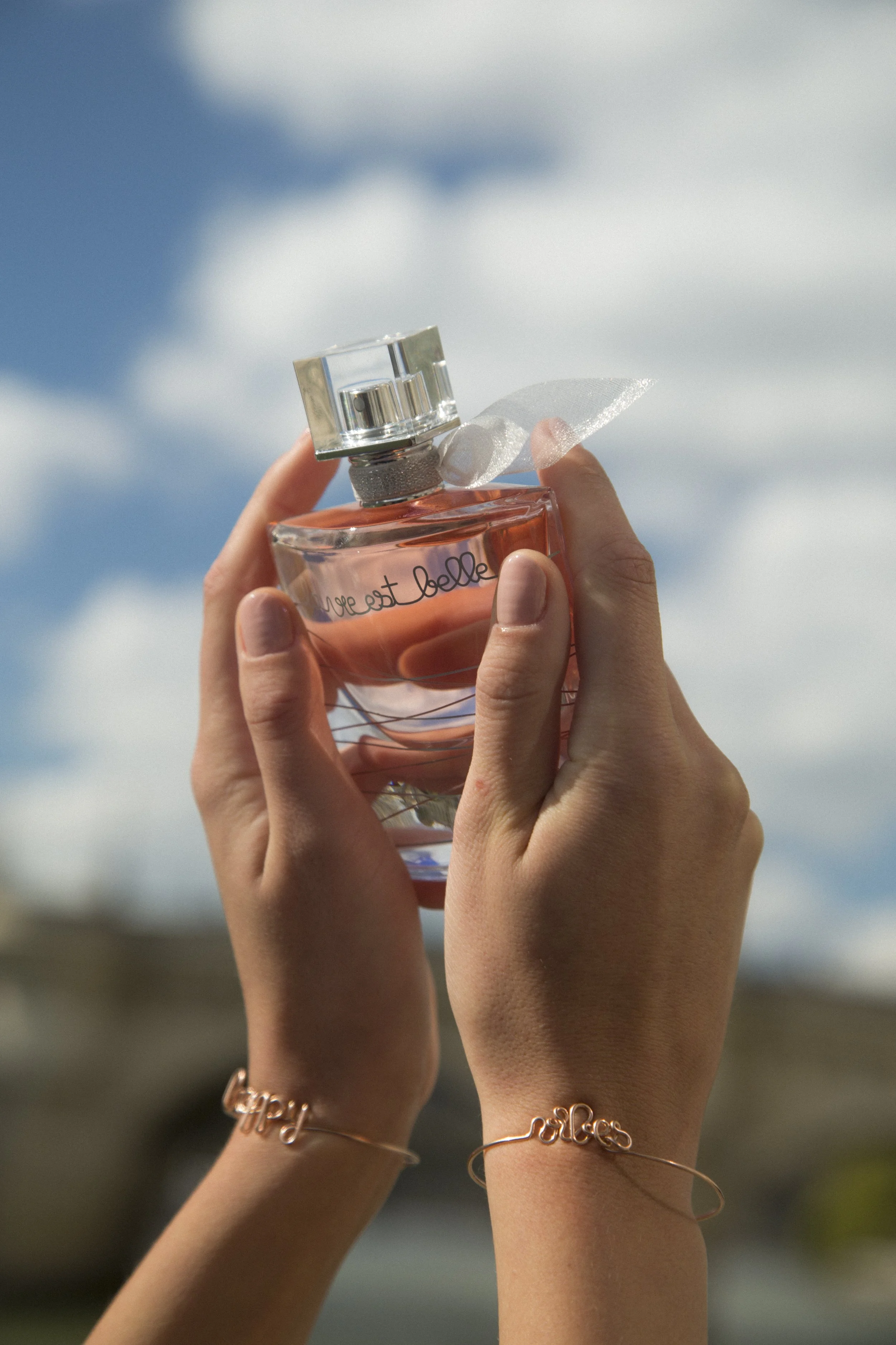 Des mains tiennent un flacon de parfum en verre clair avec du liquide rose, portant une étiquette avec l'écriture "Dream est belle". La personne porte deux bracelets fins en métal or rose avec des mots inscrits, et l'arrière-plan montre un ciel bleu 