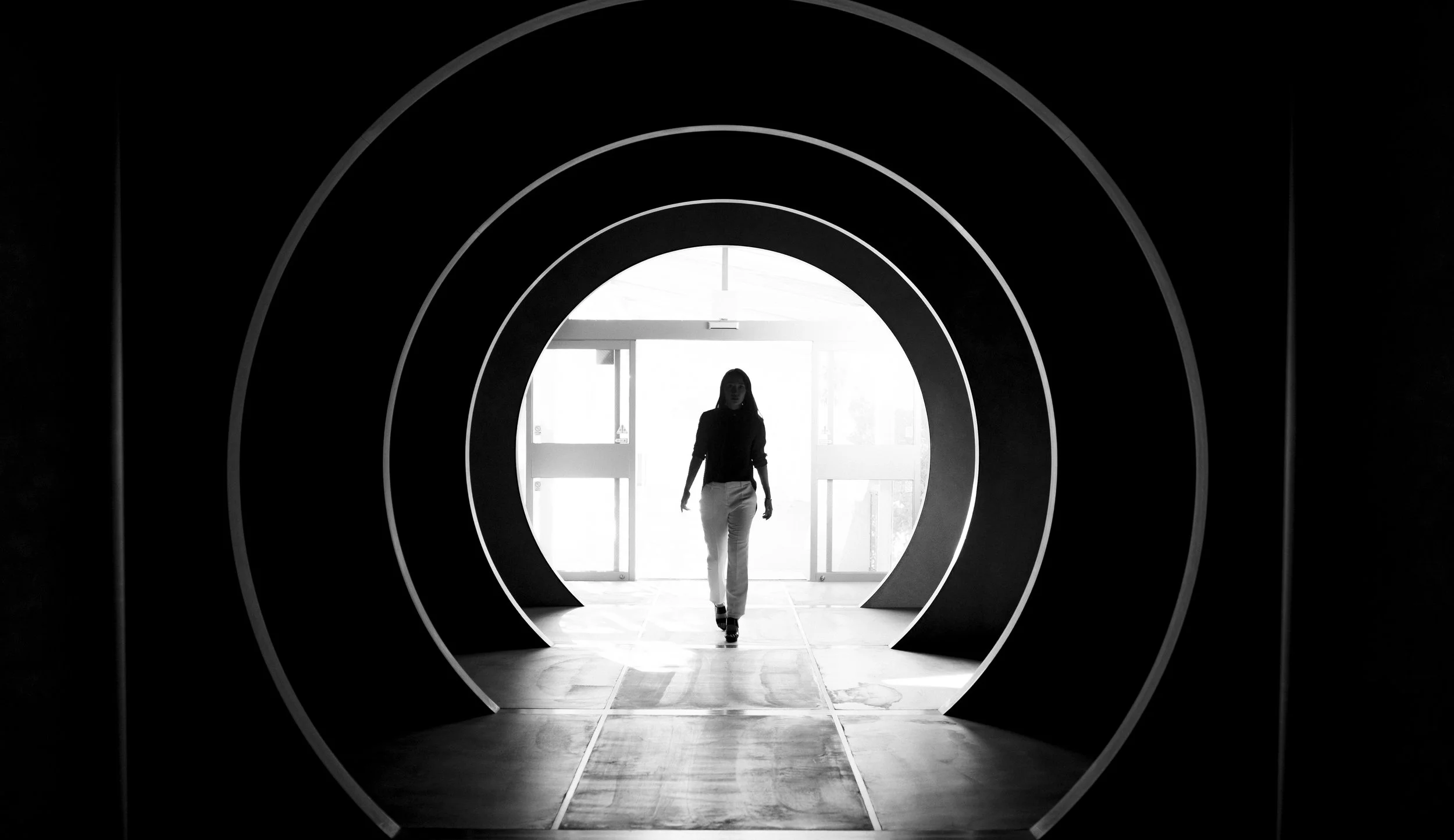 Une femme marche dans un passage circulaire sombre avec un fond lumineux, vue en silhouette.