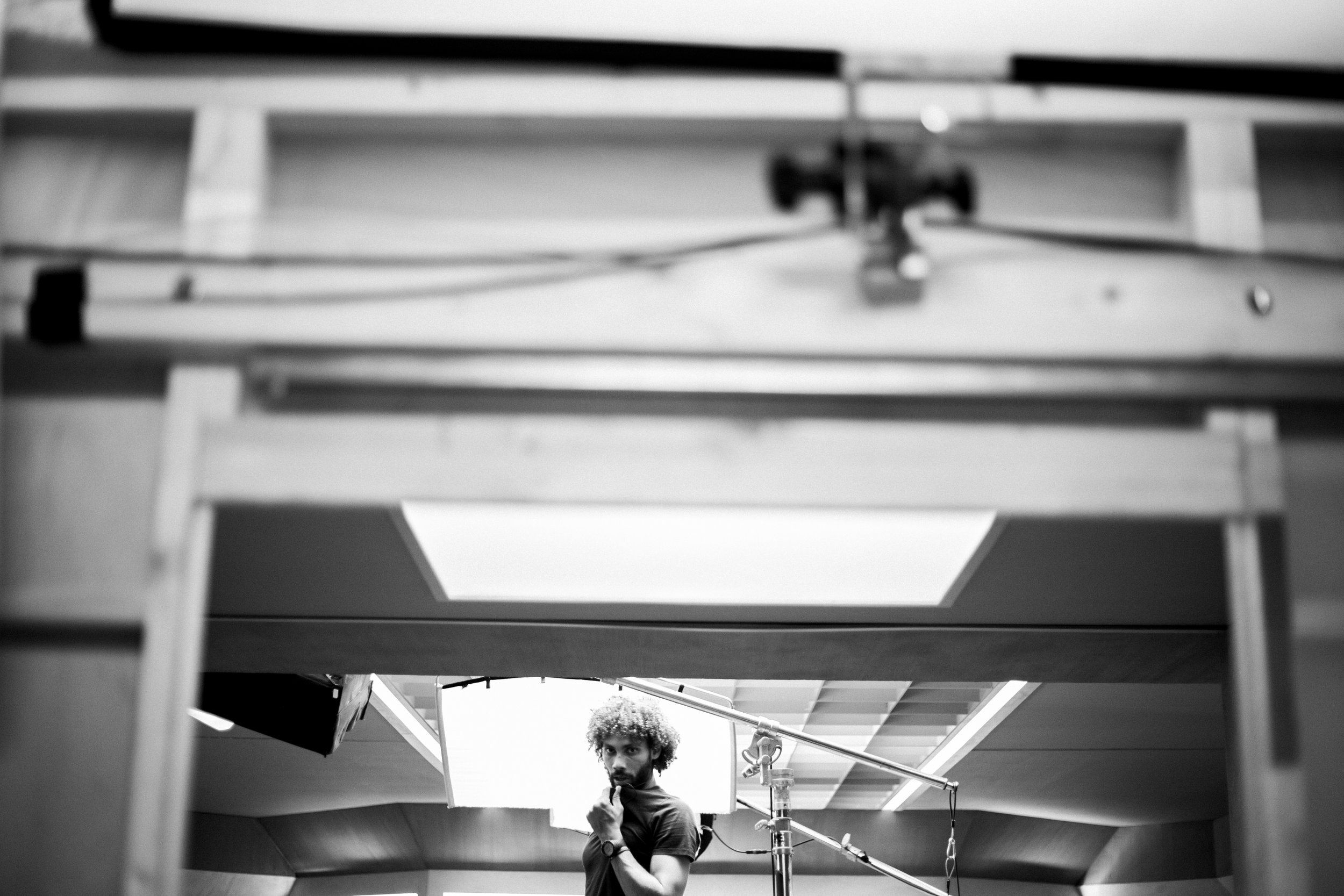 Une personne avec des cheveux bouclés regarde pensivement, derrière un écran blanc, dans une salle avec des équipements de tournage.