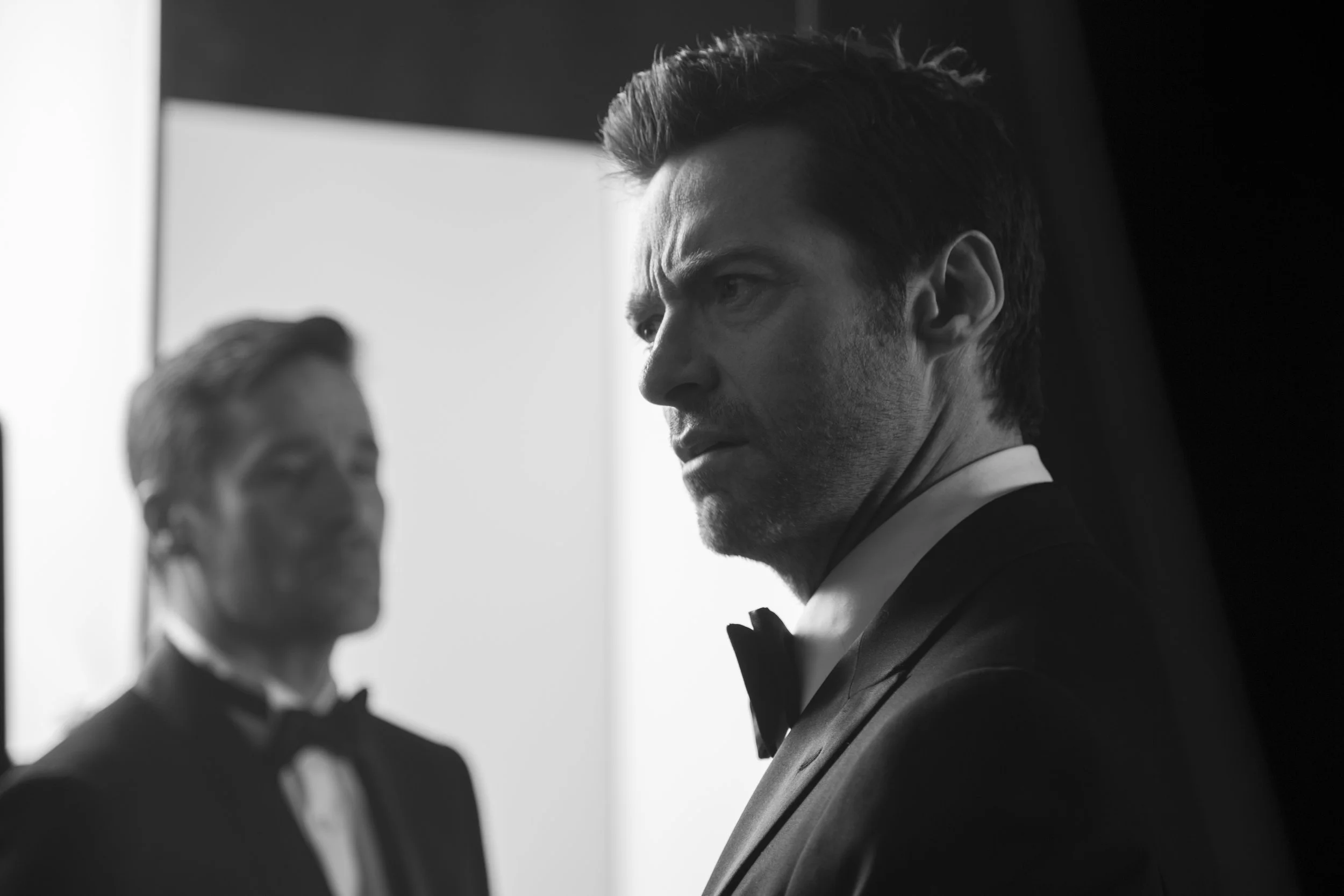 Deux hommes habillés en smoking, l'un en premier plan et l'autre flou en arrière-plan, dans une ambiance sombre, en noir et blanc.