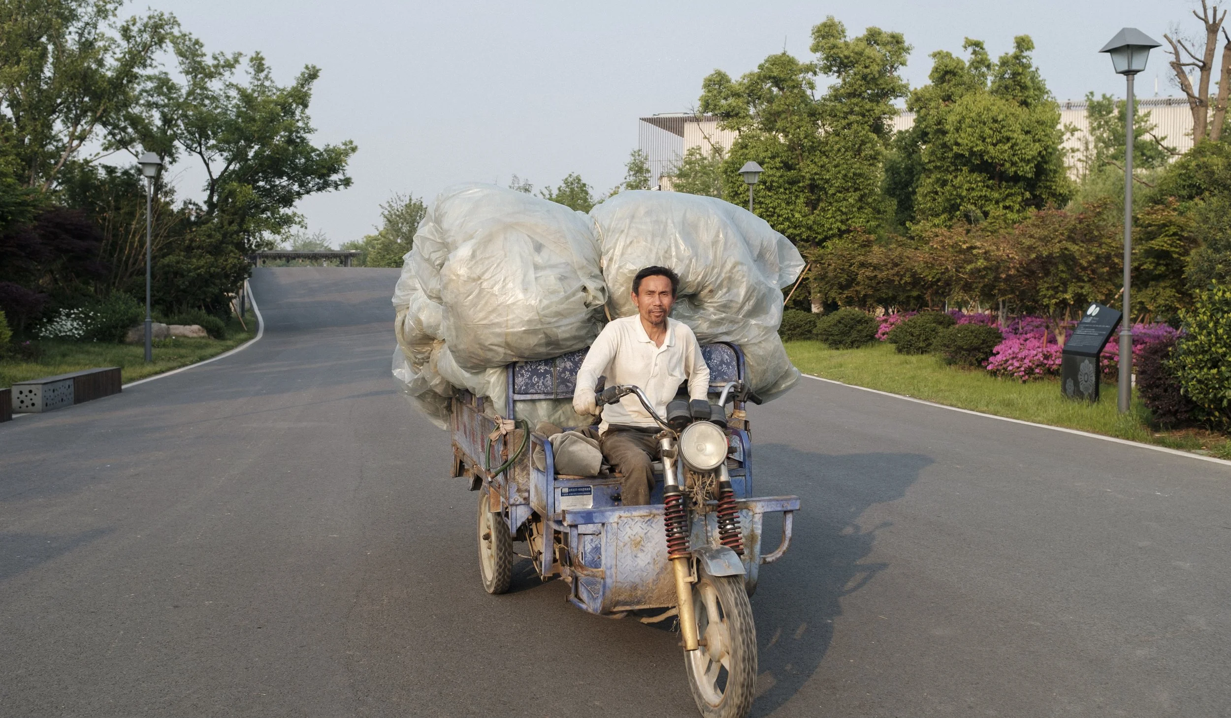 Un homme conduisant une moto-tricycle sur une route, chargé de grands sacs. Environnement verdoyant avec des arbres, des lampadaires et des fleurs.