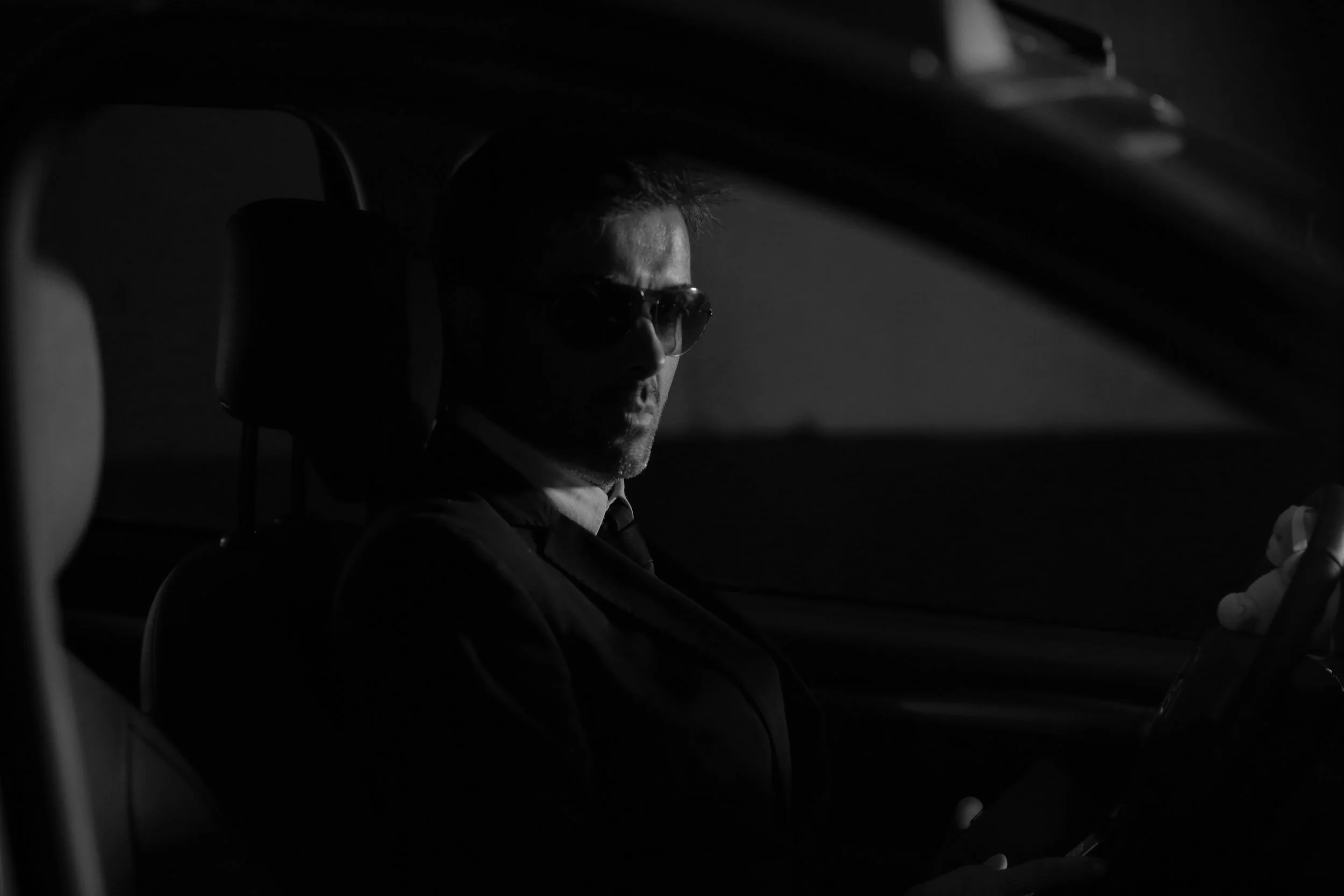 Un homme portant des lunettes de soleil, assis dans une voiture sombre.