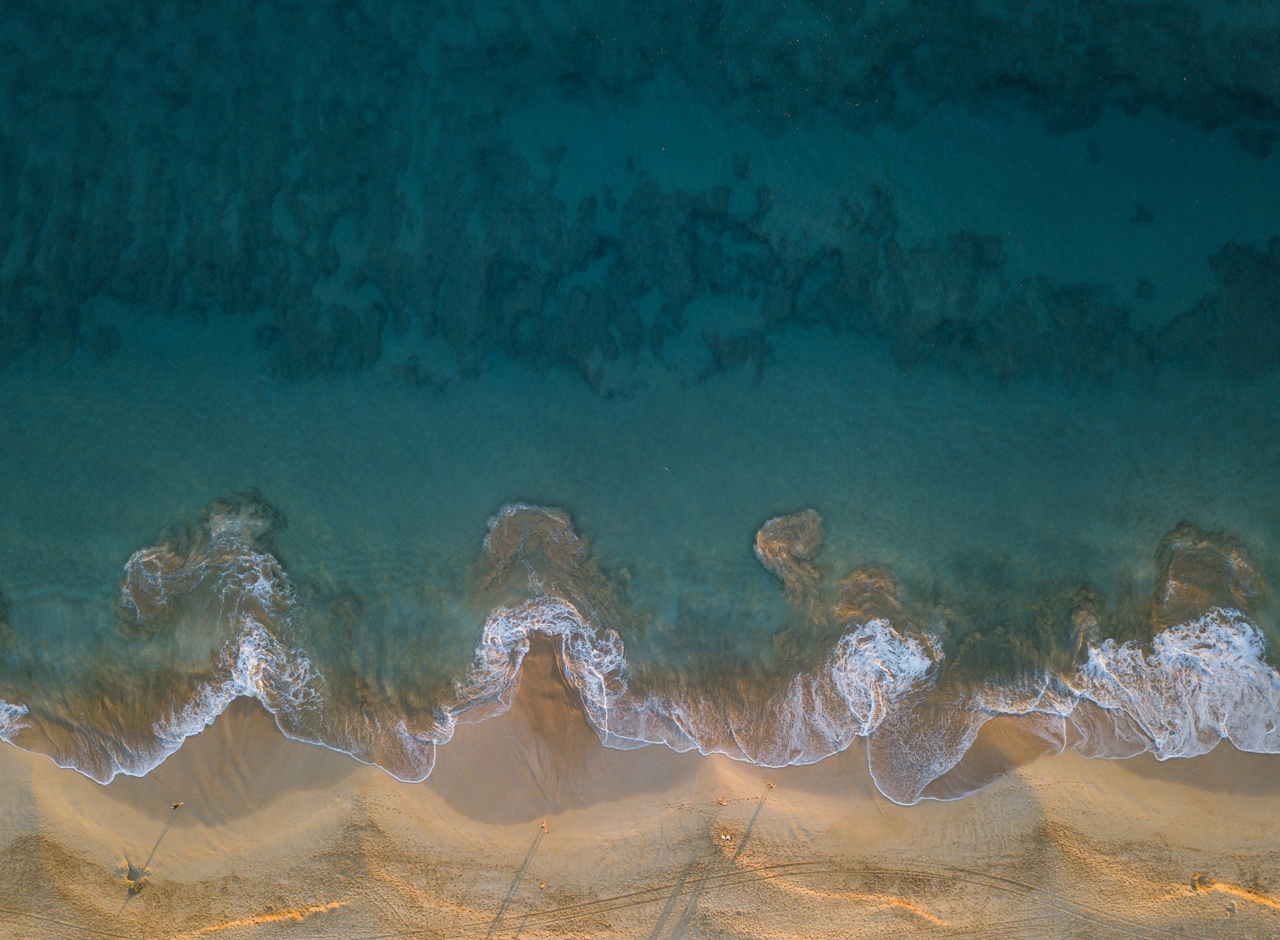 Vue aérienne d'une plage avec des vagues qui se brisent sur le sable, et quelques parasols dans le sable.