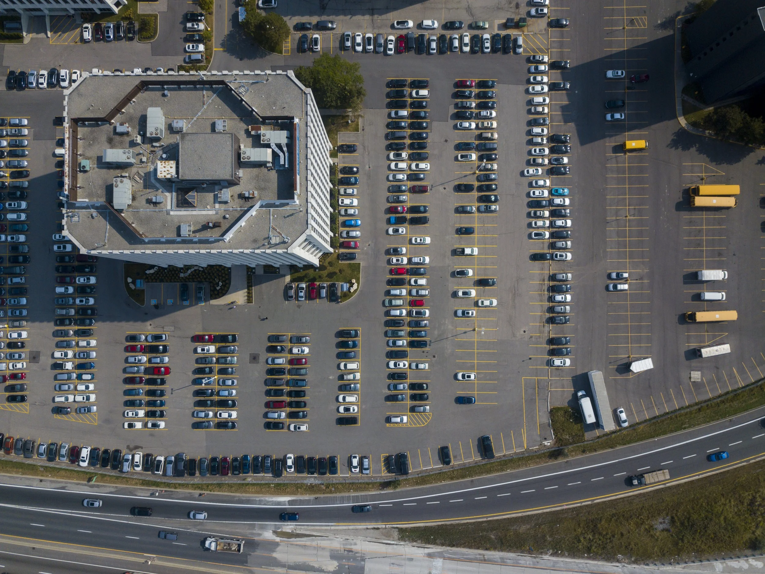 Une vue aérienne d'un parking rempli de voitures stationnées, entouré par une route située en bas de l'image et un bâtiment au centre.