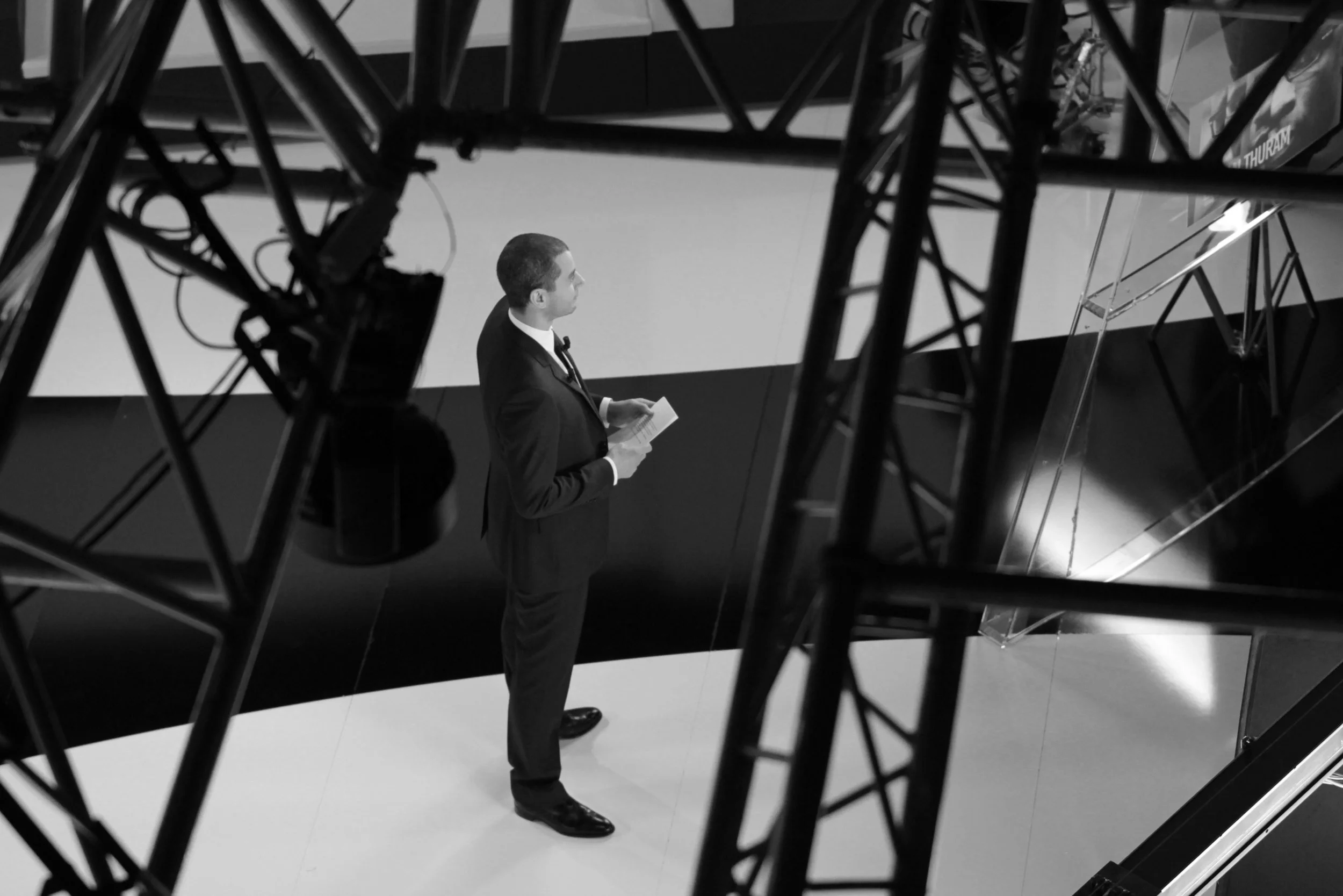 Homme en costume noir, regardant un document, vu à travers une structure métallique d'un studio ou d'une scène. Noir et blanc.