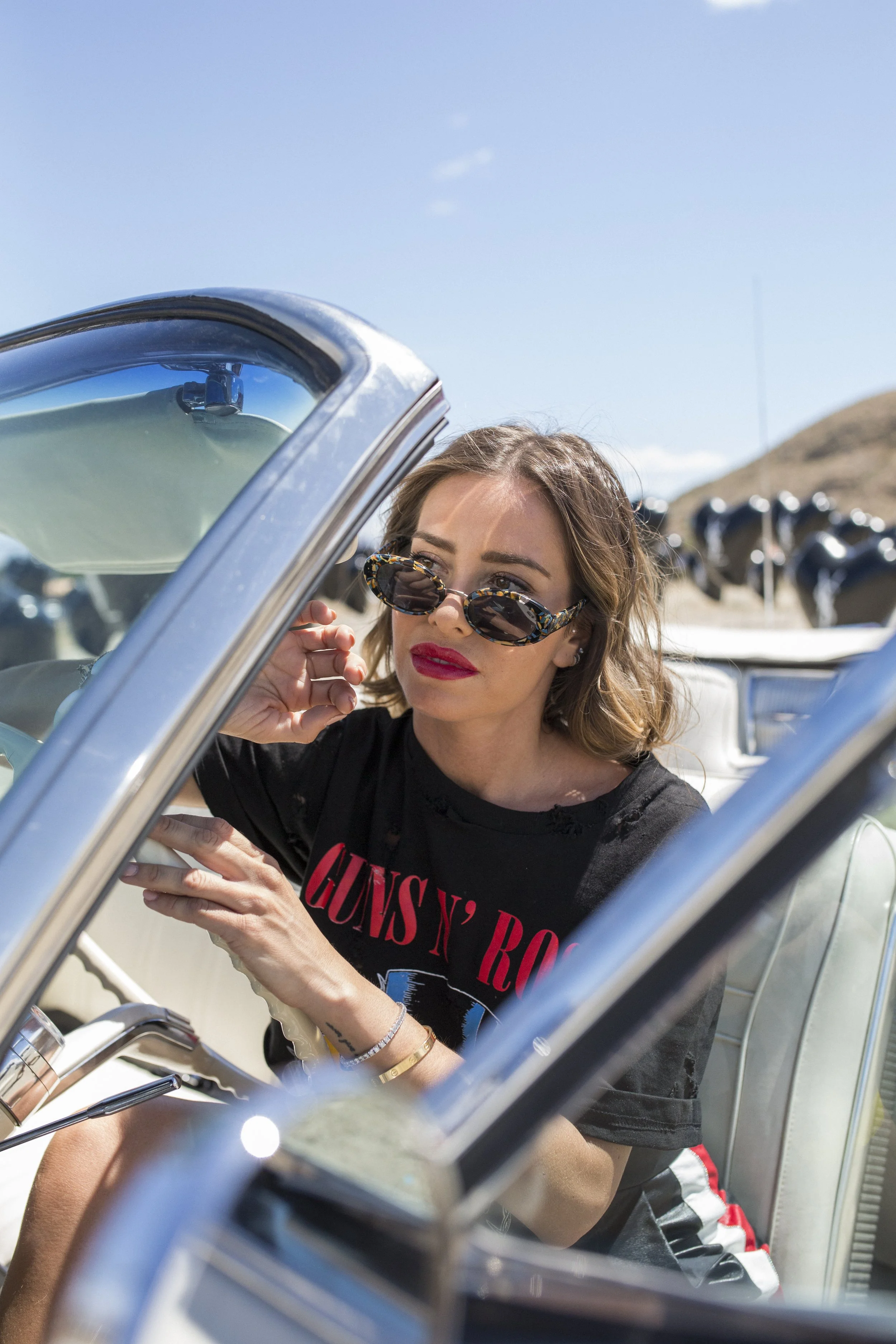 Jeune femme en lunettes de soleil, vêtue d'un t-shirt Guns N' Roses, assise dans une voiture décapotable blanche, en extérieur en journée.