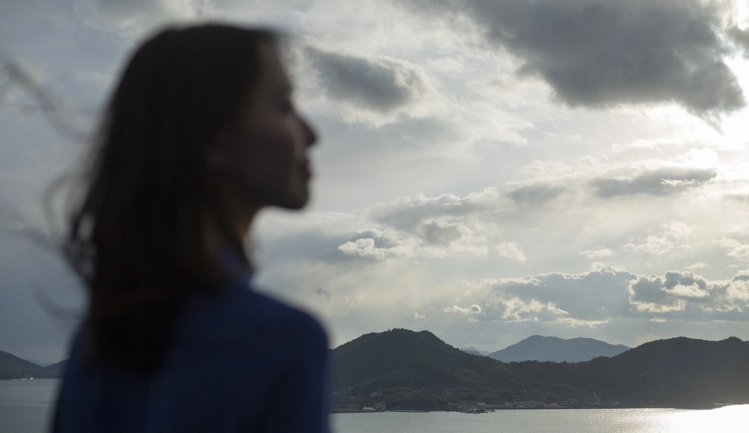 Silhouette d'une femme regardant vers la mer et les montagnes sous un ciel nuageux.