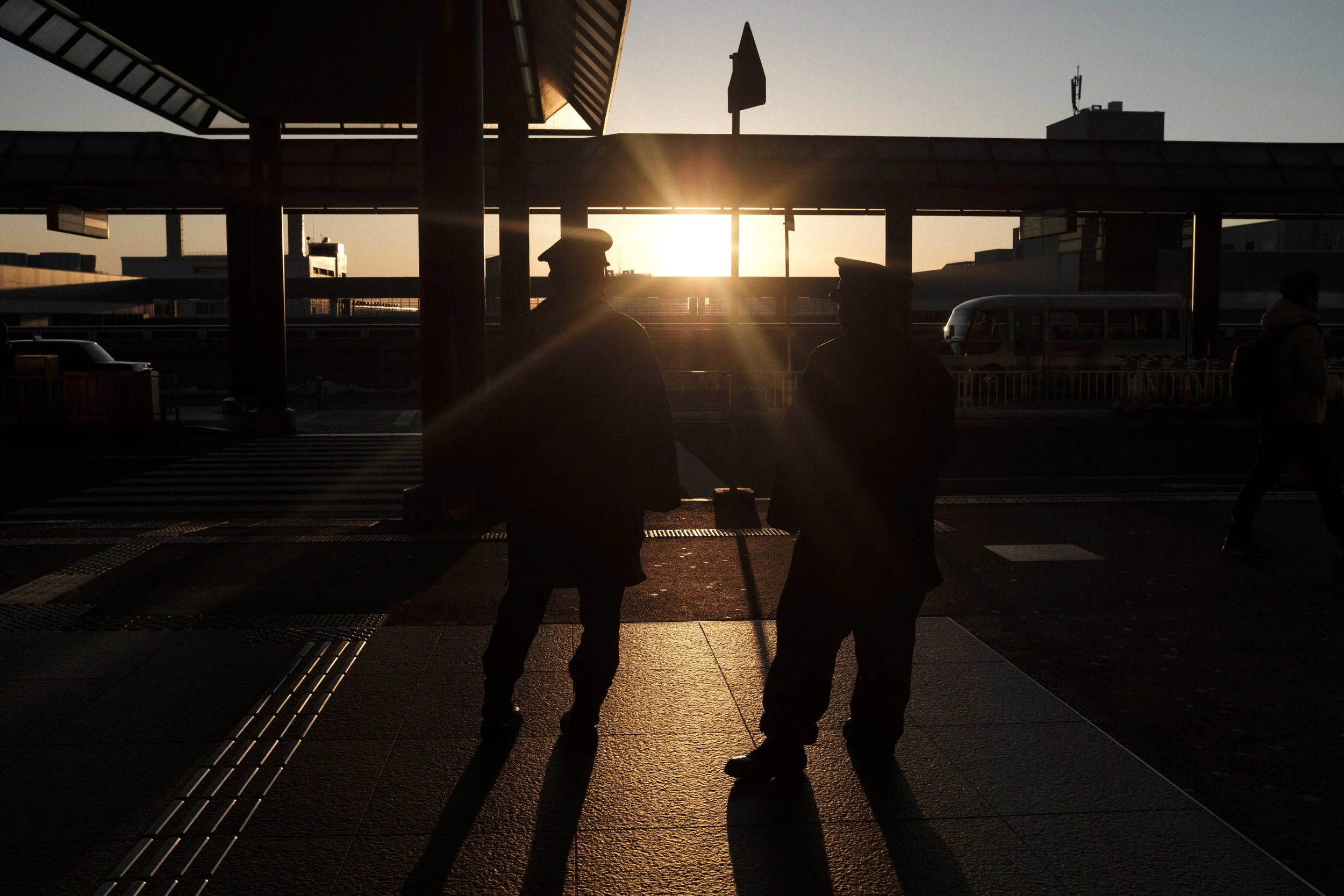 Deux hommes en uniforme de police discutent à l'extérieur d'une gare au coucher du soleil, avec des véhicules stationnés en arrière-plan.