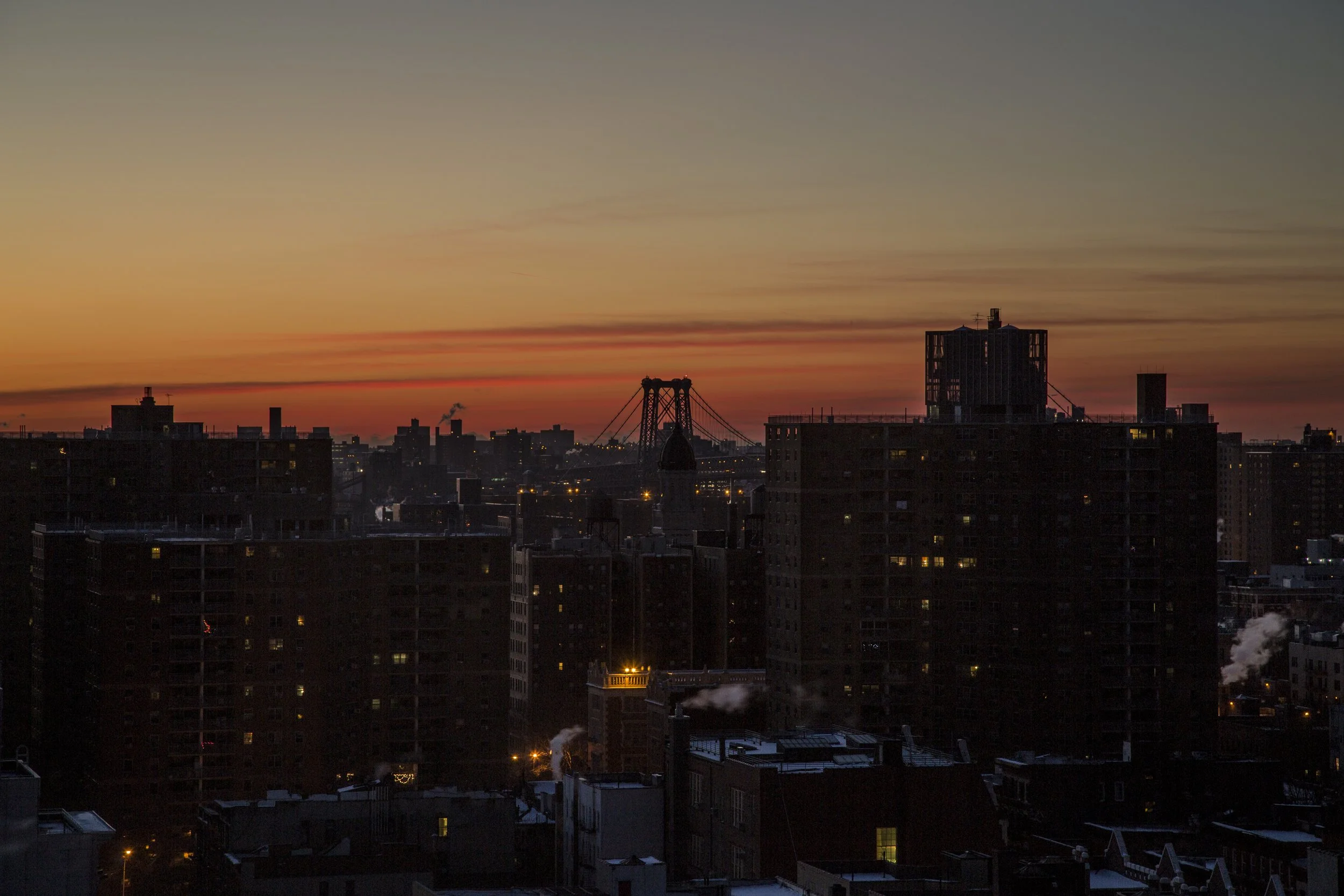 Vue sur une ville au crépuscule avec des bâtiments en silhouette et un ciel coloré d'orange et de bleu.