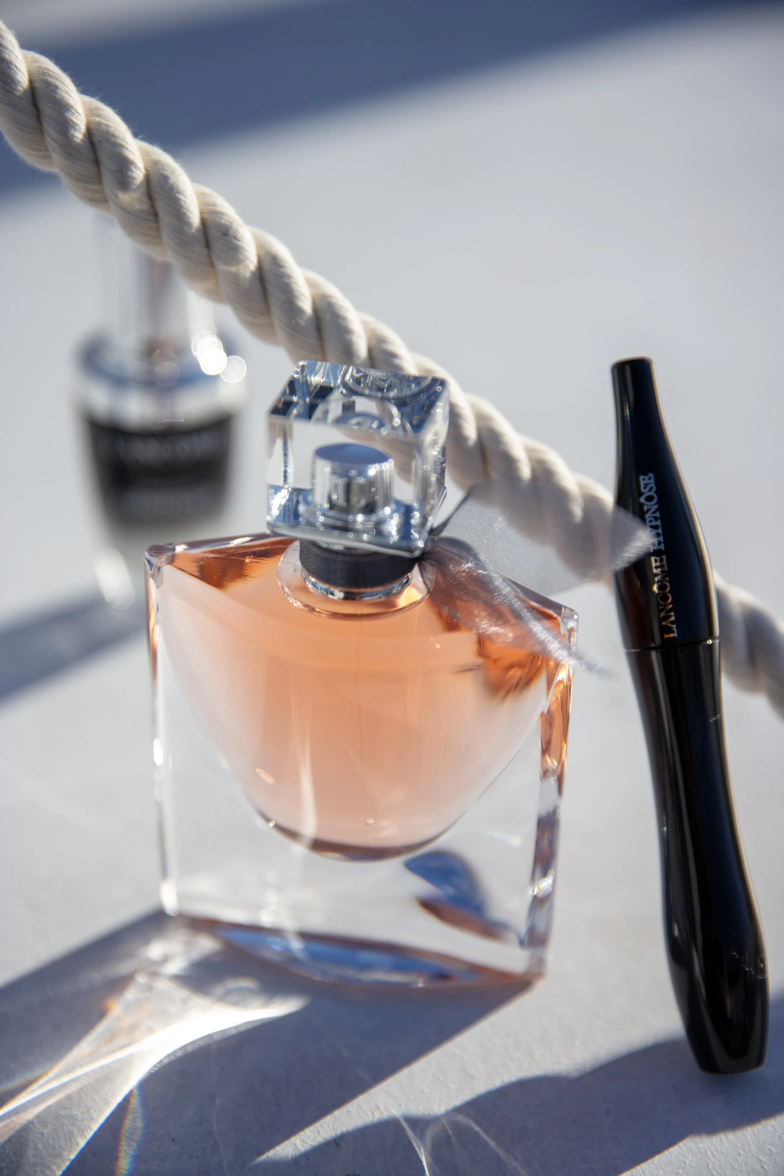 Une bouteille de parfum en verre transparent avec un liquide rose pâle, un flacon de parfum avec un bouchon en cristal, et un mascara noir avec le nom "Lancôme Hypnôse" écrit dessus, reposant sur une corde enroulée sur une surface blanche sous une lu