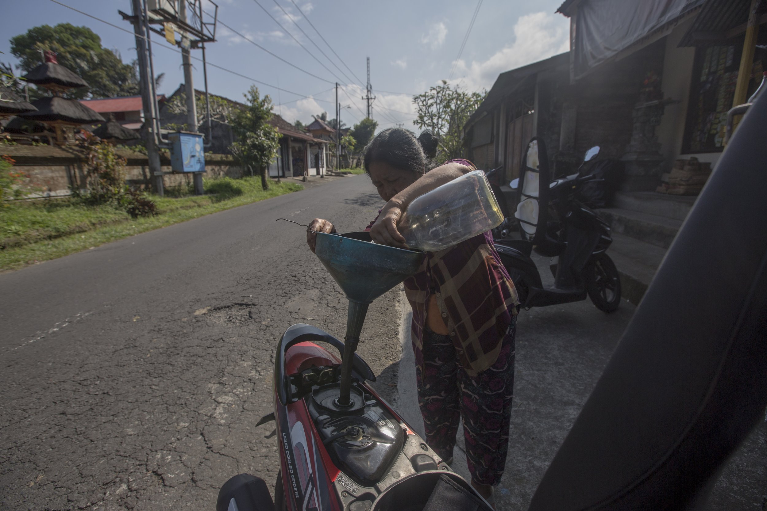 Une femme âgée remplit un verre à partir d'une fontaine à eau installée sur une moto dans une rue de village, avec des maisons traditionnelles en arrière-plan.