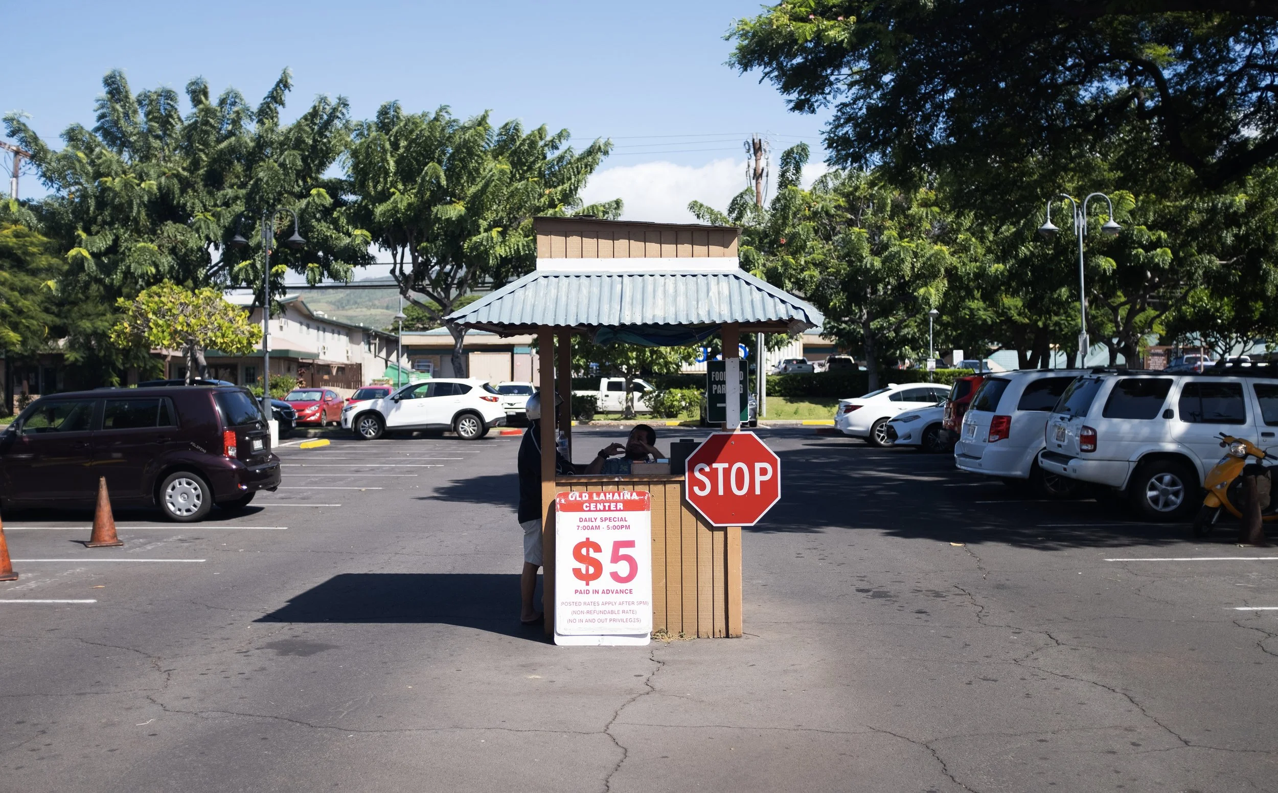 Un poste de contrôle de parking avec un arrêt STOP, une tige de paiement de $5, et deux personnes en train de discuter derrière le comptoir, dans un parking en plein jour avec des voitures stationnées et des arbres en arrière-plan.