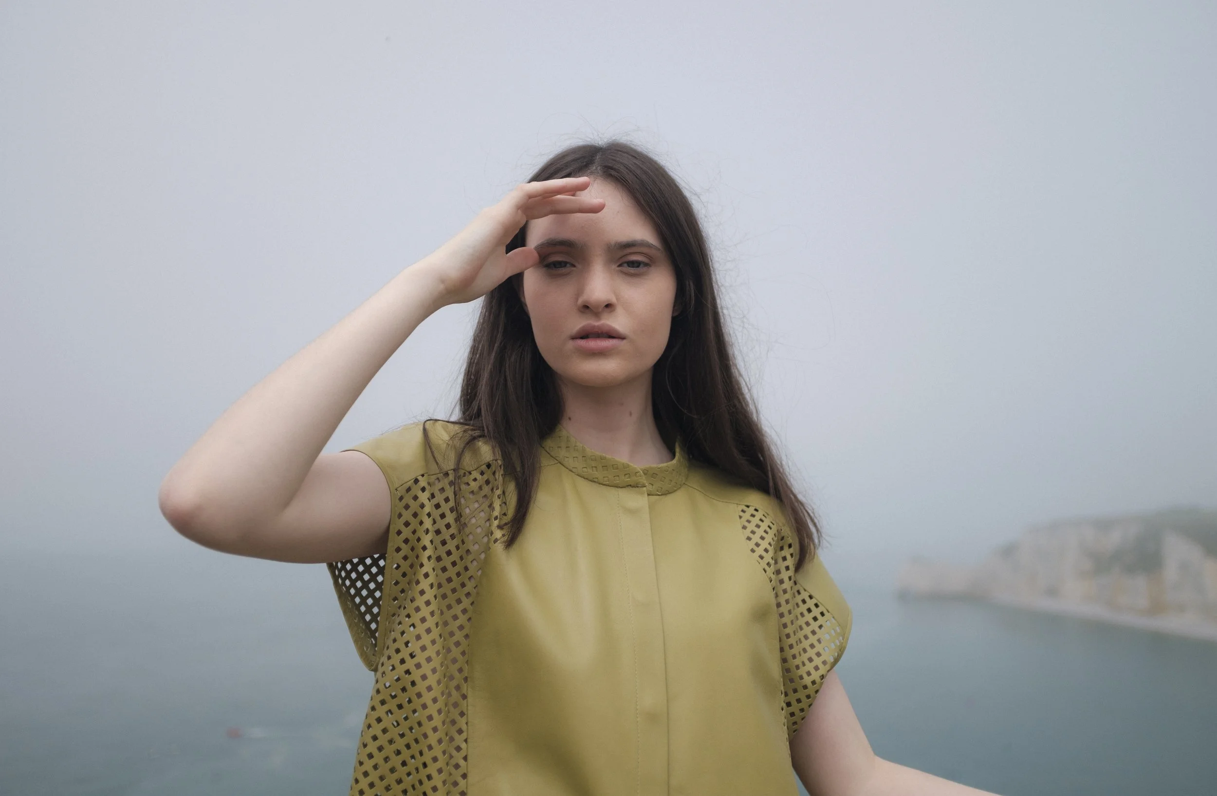 Jeune femme aux cheveux bruns, portant une robe jaune avec des motifs perforés, se tenant devant une vue brumeuse sur un corps d'eau, en train de faire un geste de salut avec la main gauche