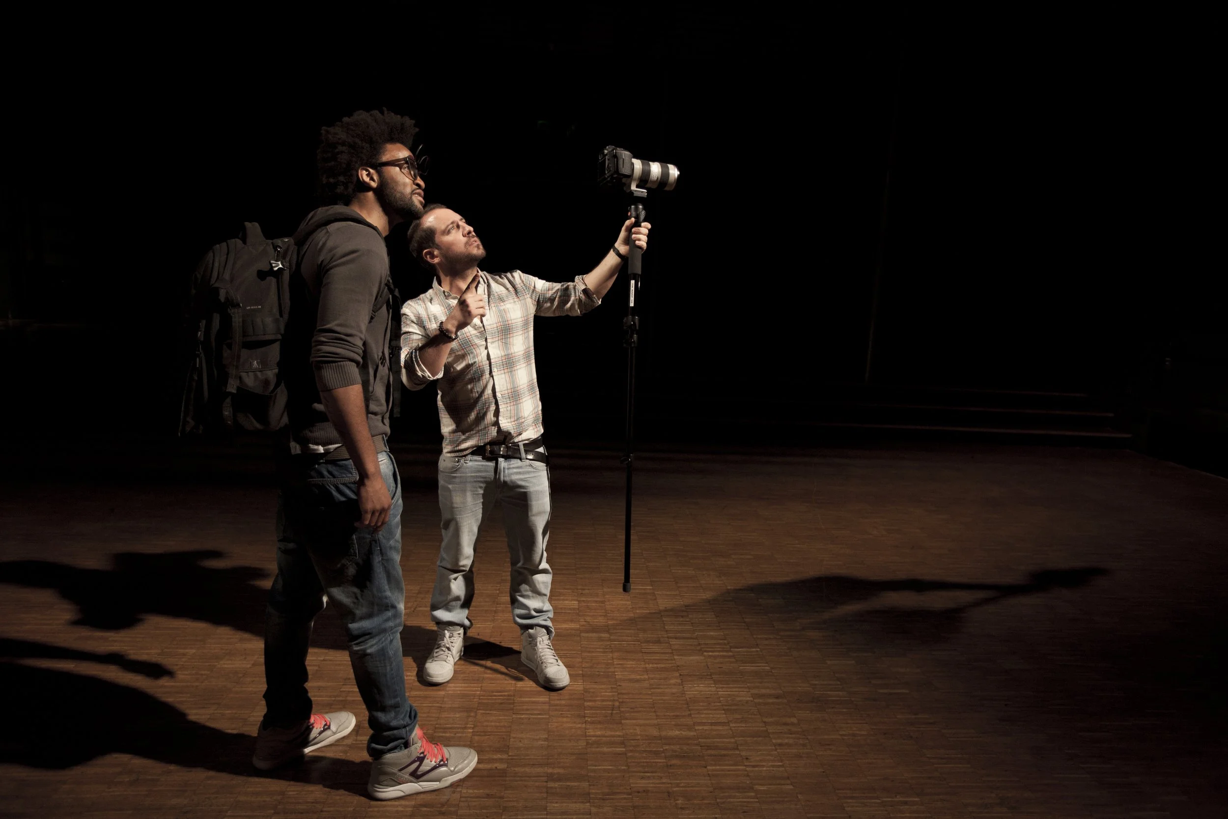 Deux hommes, l'un avec un sac à dos et l'autre pointant une caméra vers l'avant dans une pièce sombre.