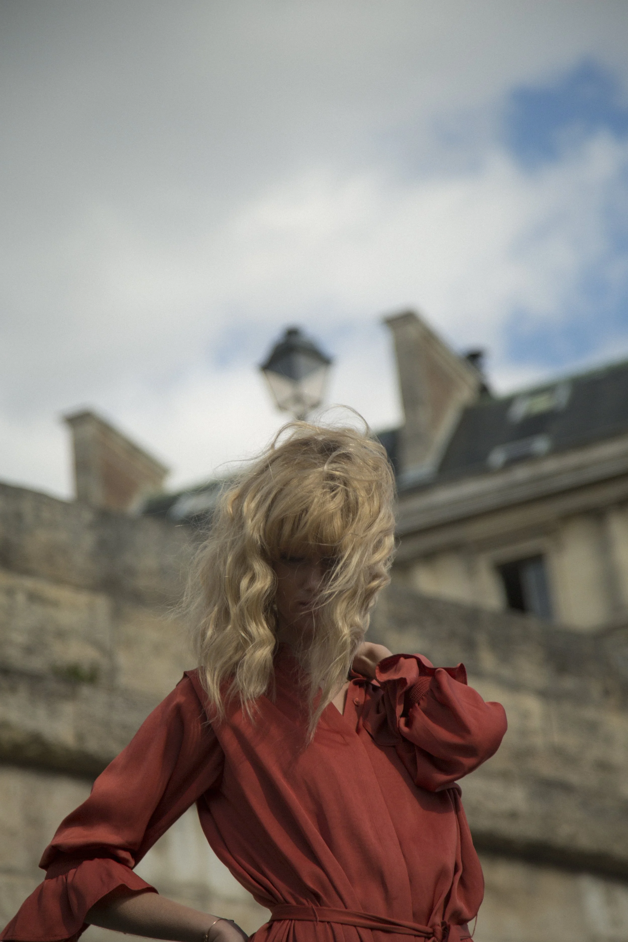 Une femme aux cheveux longs et bouclés, portant une robe rouge, se tient devant un bâtiment ancien avec un ciel nuageux en arrière-plan.