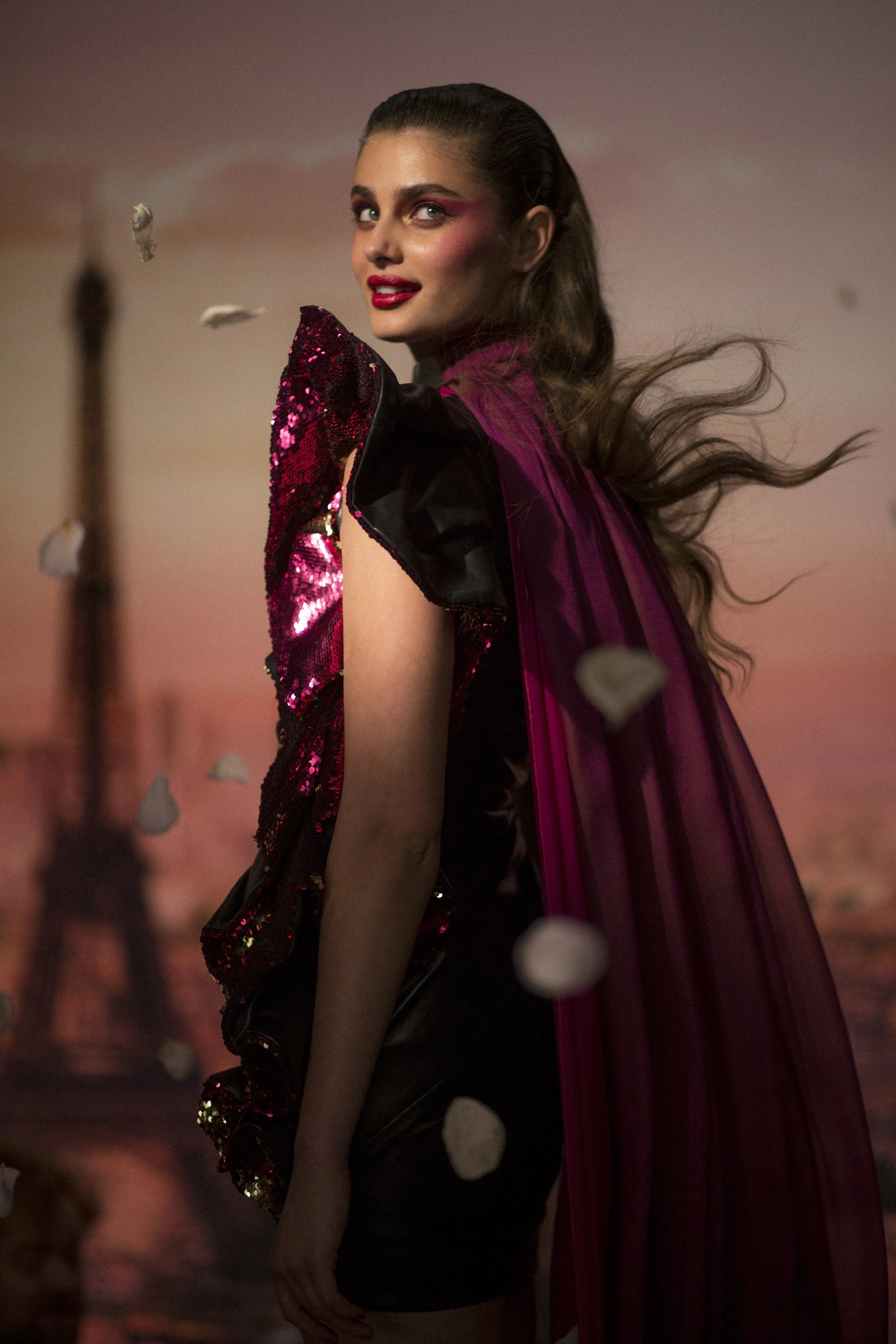 Une femme habillée d'une tenue brillante noire avec un cape rose foncé, devant un coucher de soleil avec la Tour Eiffel en arrière-plan, et des pétales qui tombent autour d'elle.