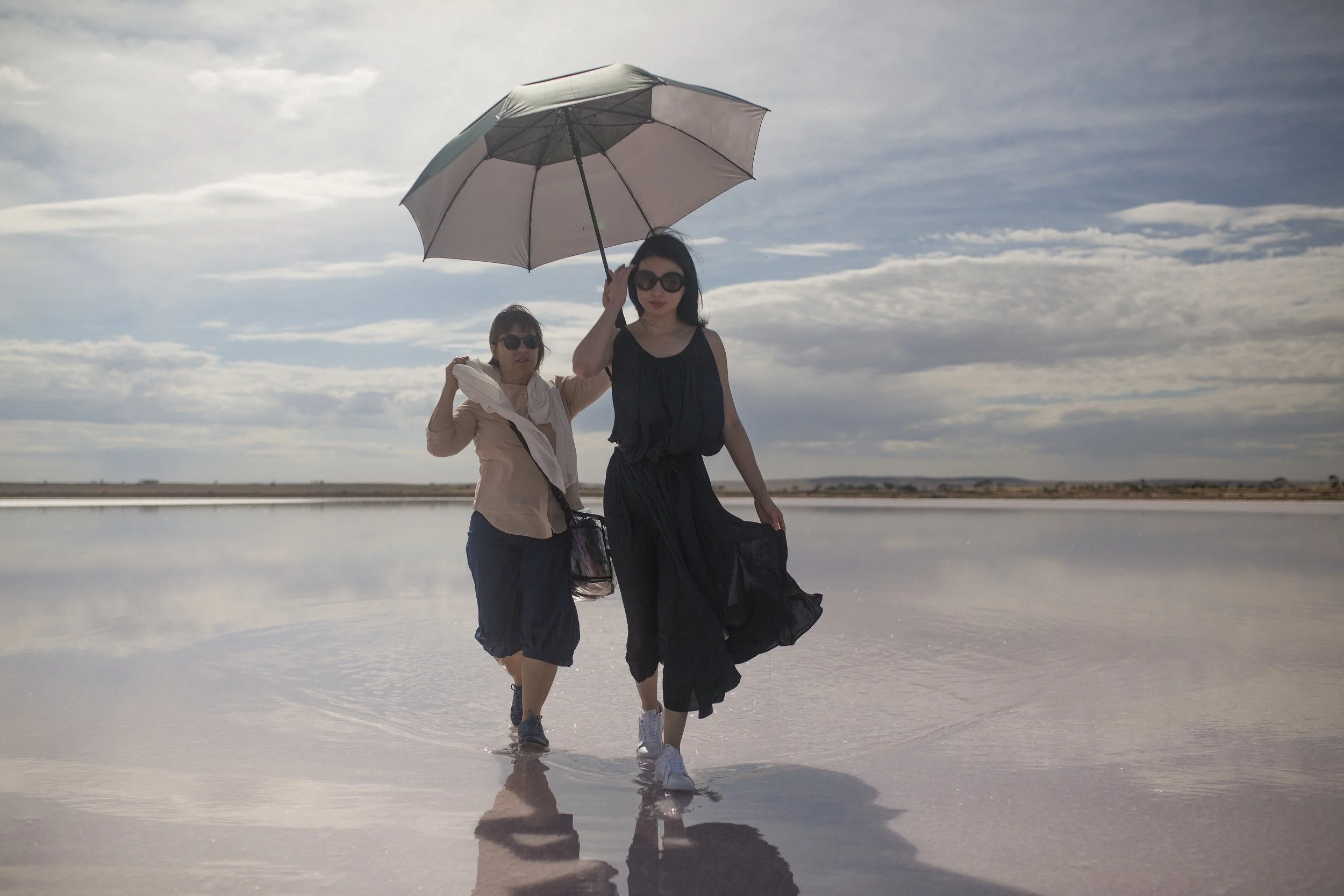 Deux femmes marchent dans une zone humide avec un ciel nuageux, une d'elles tient un parapluie beige. La surface de l'eau reflète le paysage et le ciel.