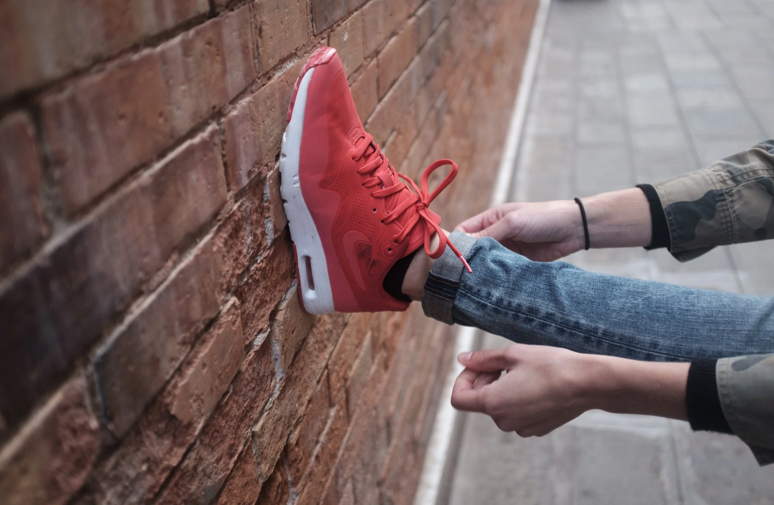 Une personne portant une chaussure de course rouge, qui repose contre un mur en briques, en train de lacer ou de détacher la chaussure.