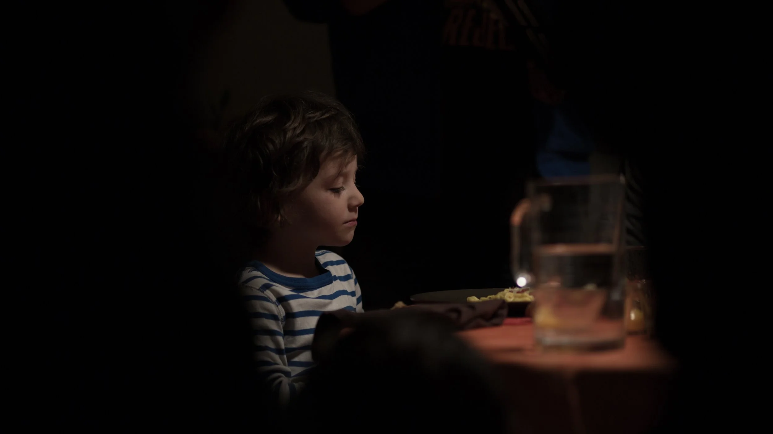 Un jeune garçon avec les yeux fermés, assis à une table dans une pièce sombre, éclairée par une lumière chaude, avec un verre d'eau et une assiette de nourriture devant lui.