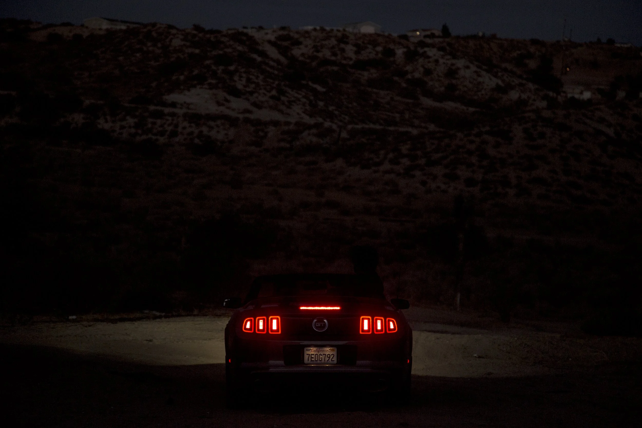 Voiture noire avec feux arrière allumés, vue de derrière, dans un paysage nocturne avec des collines en arrière-plan.