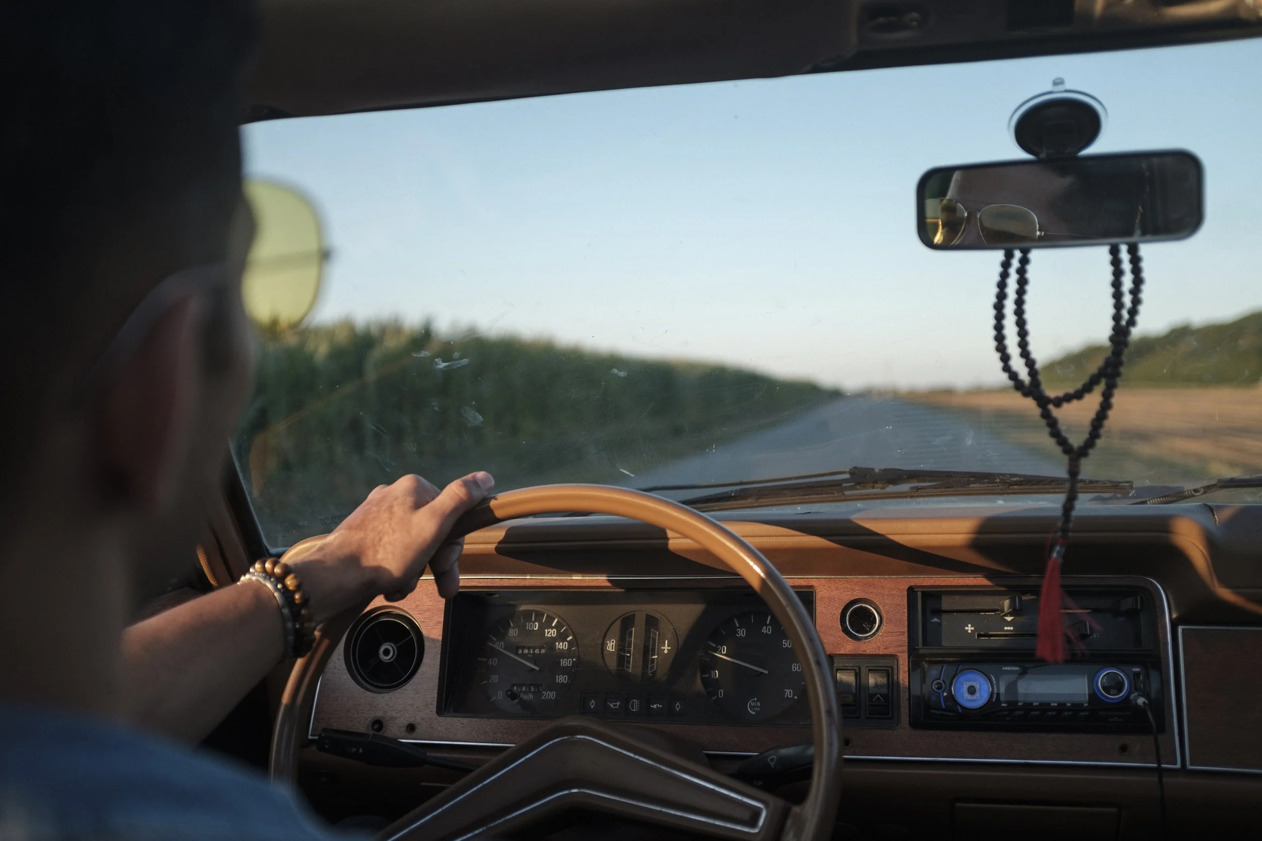 Vue depuis l'intérieur d'un véhicule classique, montrant le conducteur avec une main sur le volant, la route et le paysage vert à l'extérieur, avec un tableau de bord vintage et un miroir avec un pendentif de perles suspendu.